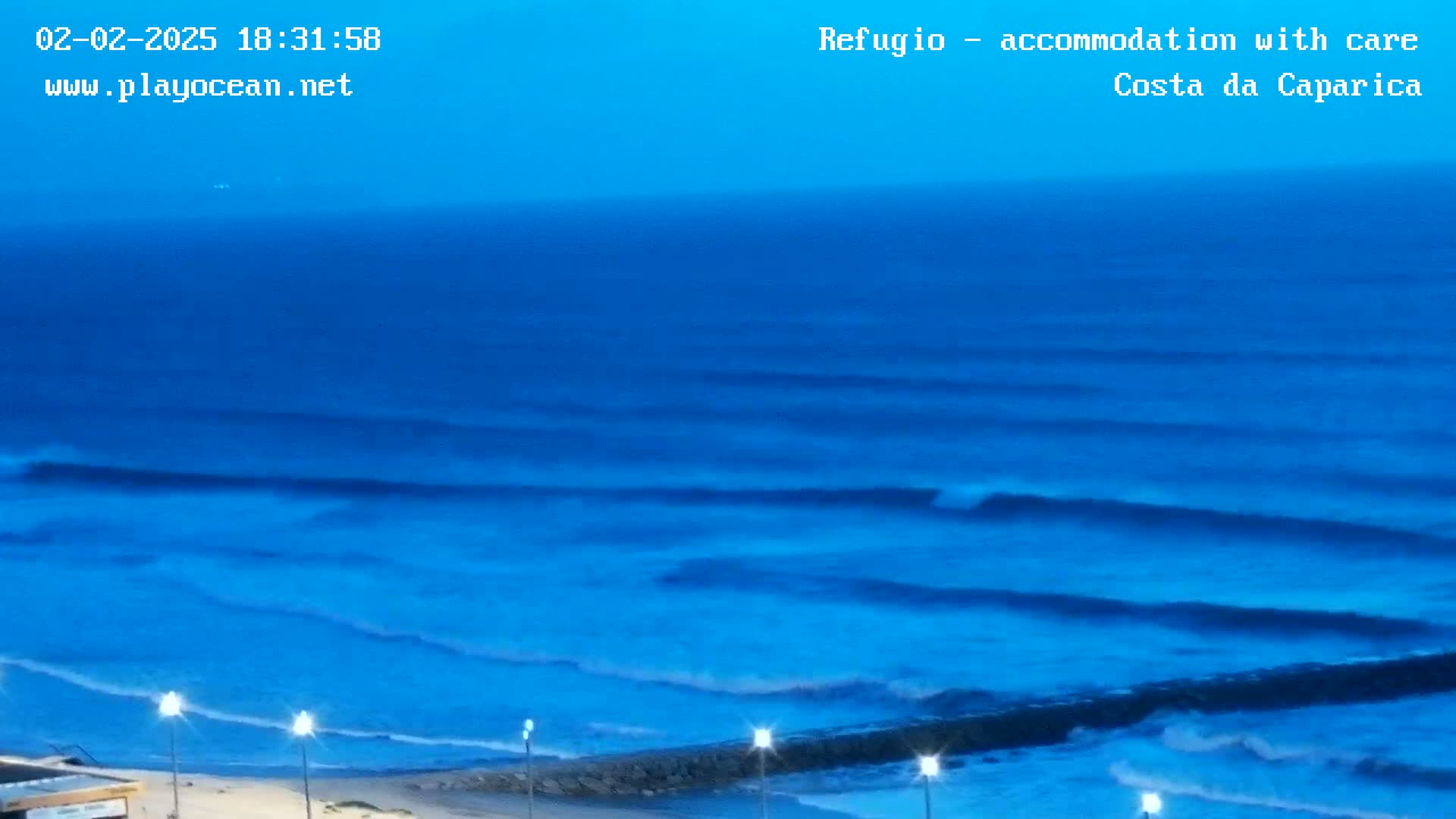 A twilight view of ocean waves rolling onto a shore with a low wall and lights along the beach.