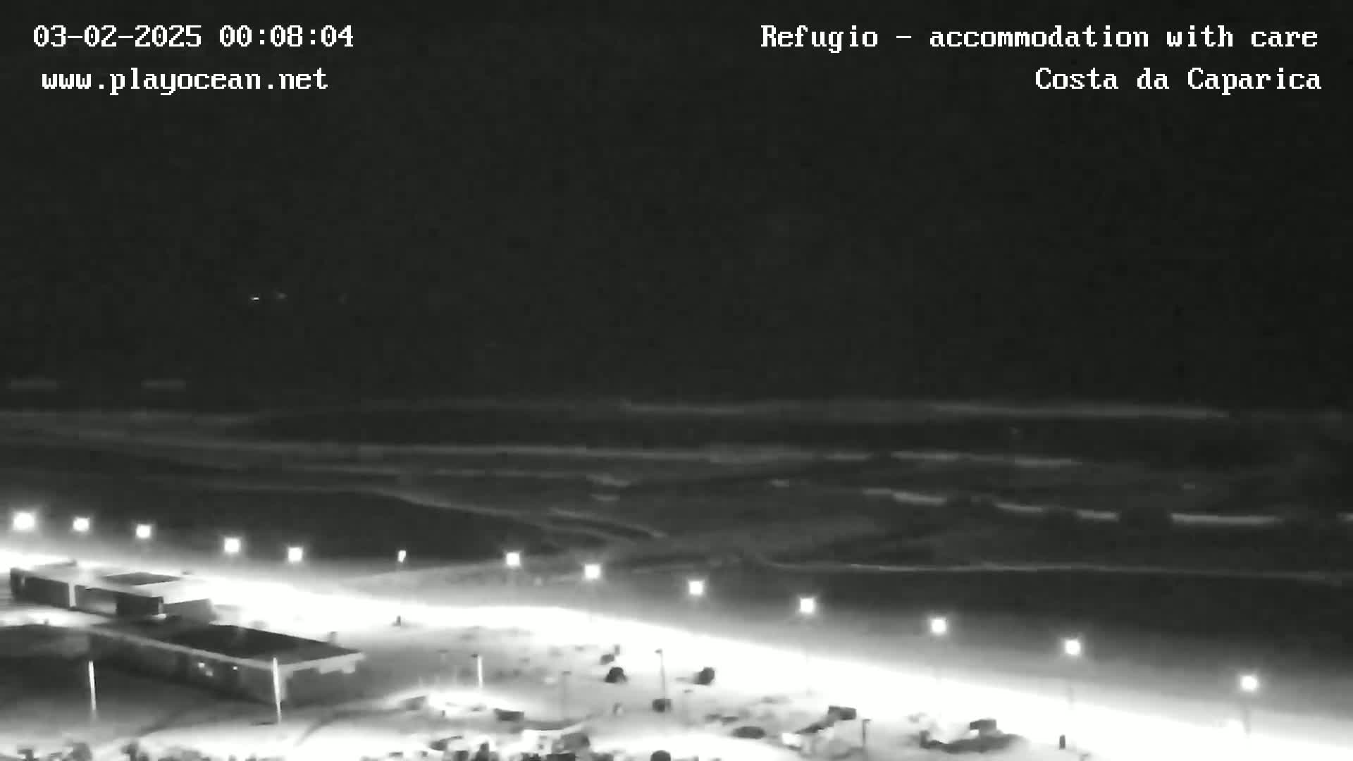 A nighttime, low-light view shows a dark beach with calm waves under a clear sky, illuminated by streetlights lining a road near buildings.