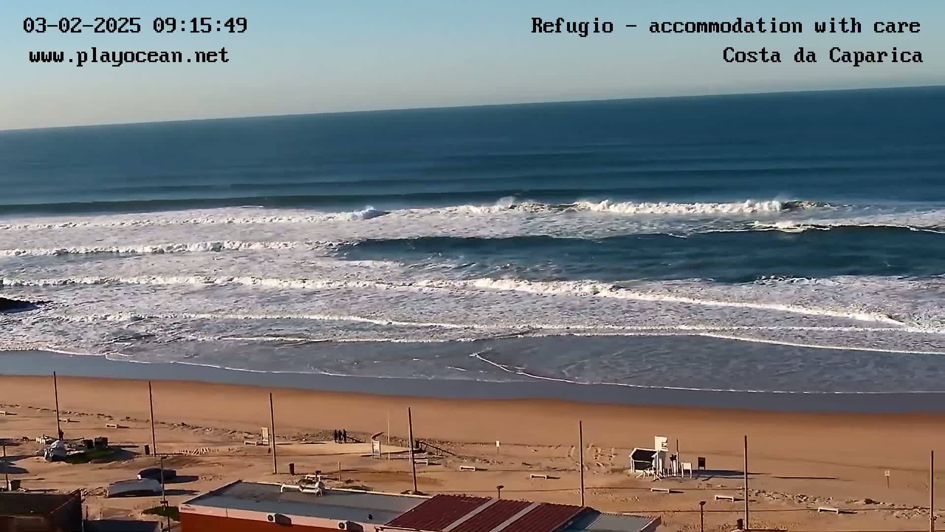 A wide shot shows a sandy beach with moderate waves rolling in under a clear, sunny sky.