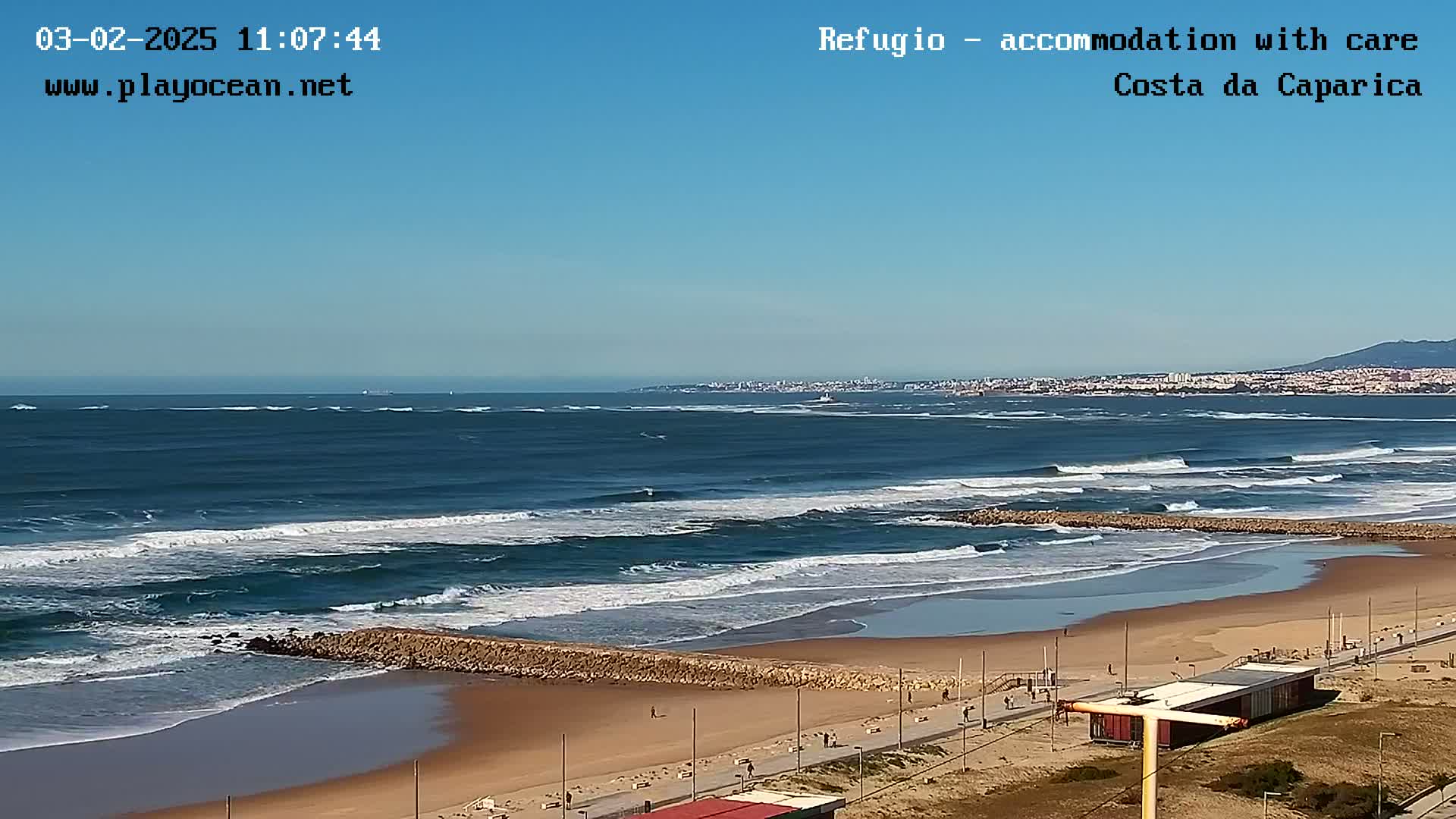 An ocean beach with a clear blue sky shows moderate waves breaking on a sandy shore with a paved walkway and a few small buildings in the distance.