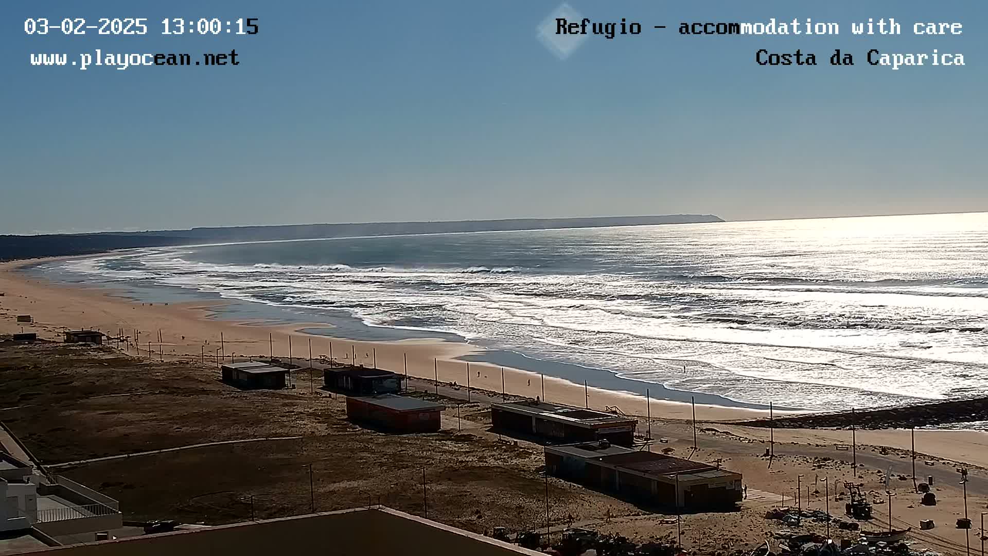 A sandy beach with small buildings stretches along a coastline, where moderate waves roll in under a bright, sunny sky.