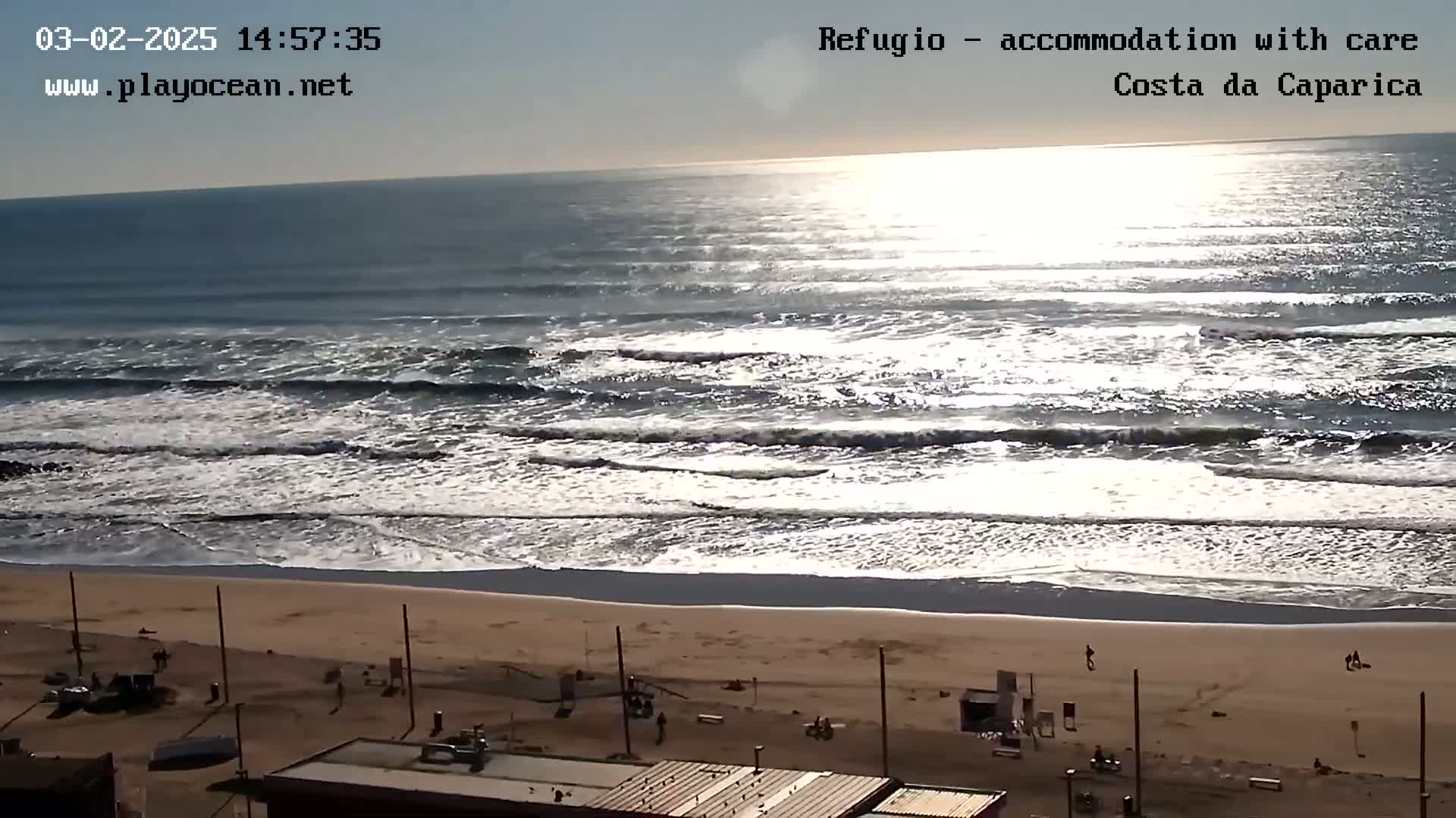 A sunny day at a beach shows waves rolling onto a sandy shore with a few people visible on the beach and buildings in the foreground.
