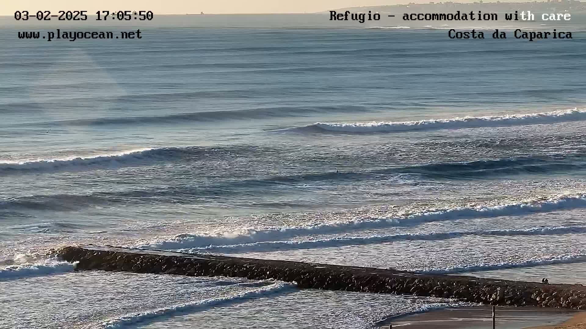A long, dark jetty extends into a calm ocean with several lines of gently rolling waves under a clear, bright sky.