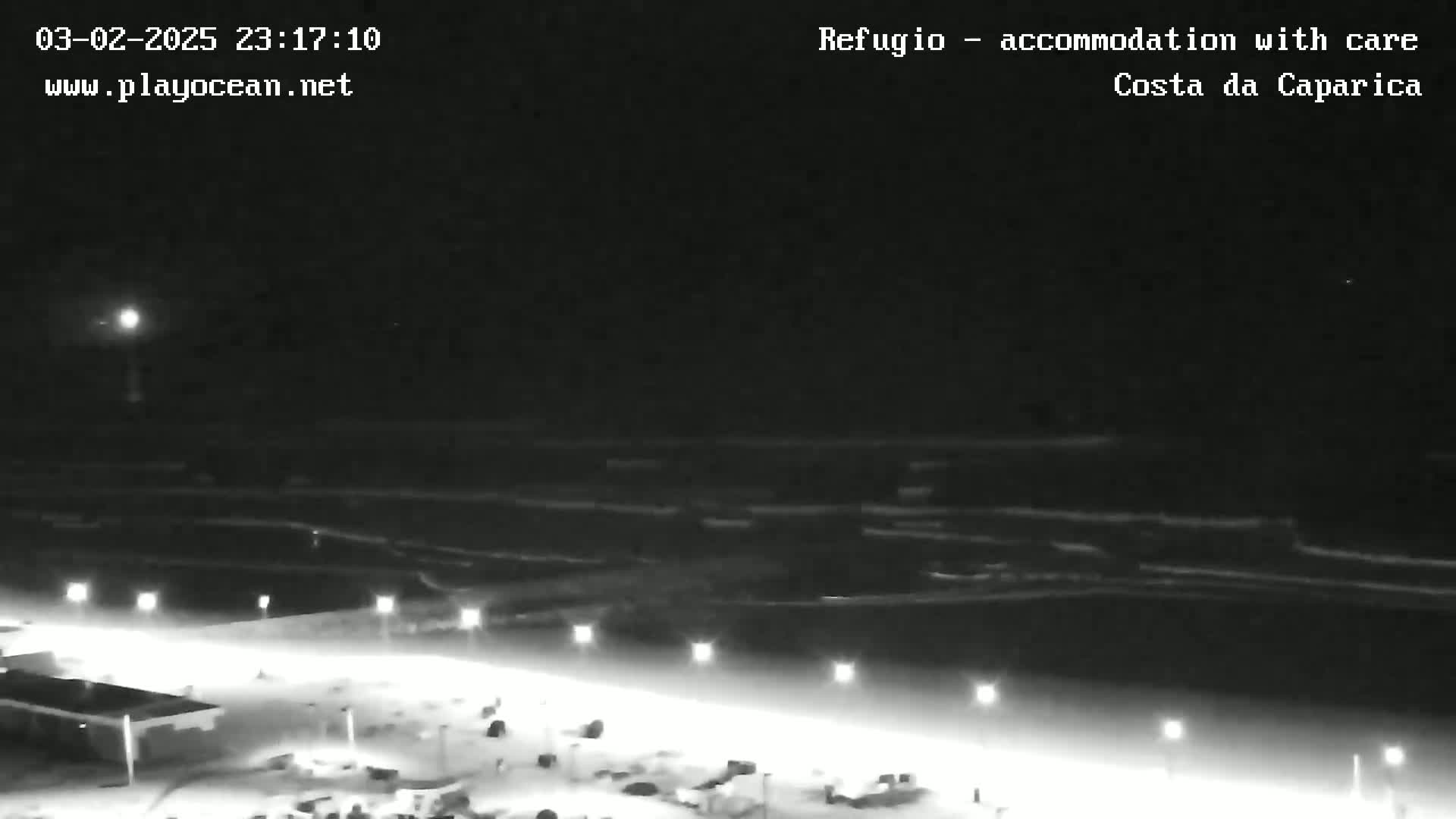 A nighttime, low-light view of a beach with a dark ocean, illuminated by several lights along a walkway.