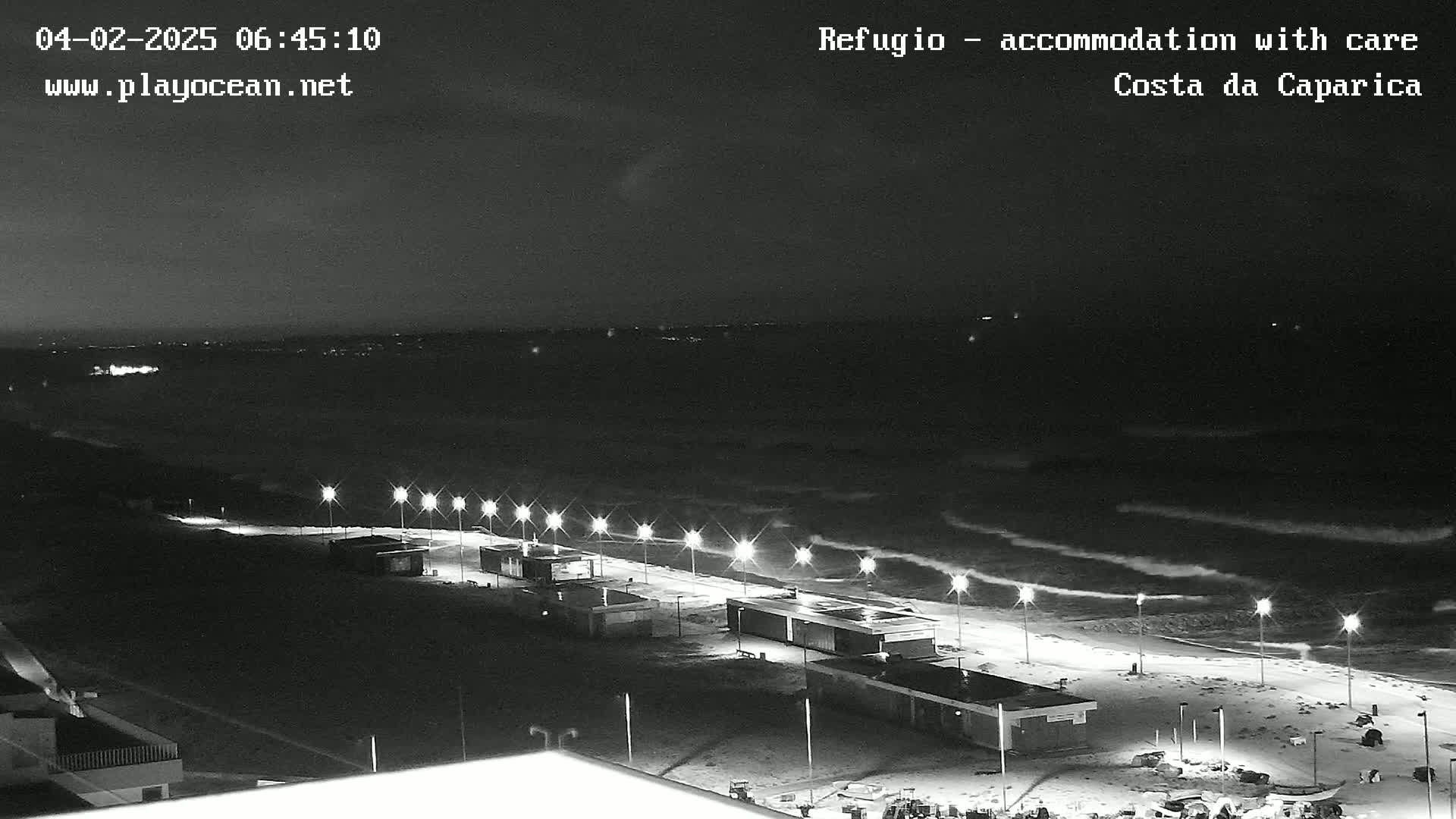 A nighttime, black and white view of a beach at night with dark, choppy waves under a mostly clear sky, illuminated by lights lining a paved walkway.