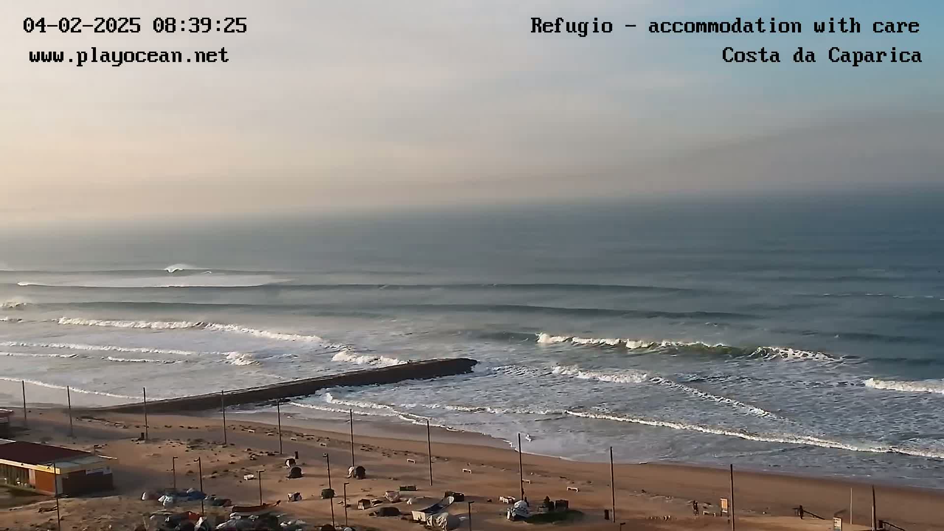 A beach with a calm ocean under a hazy sky shows moderate-sized waves rolling onto the shore.