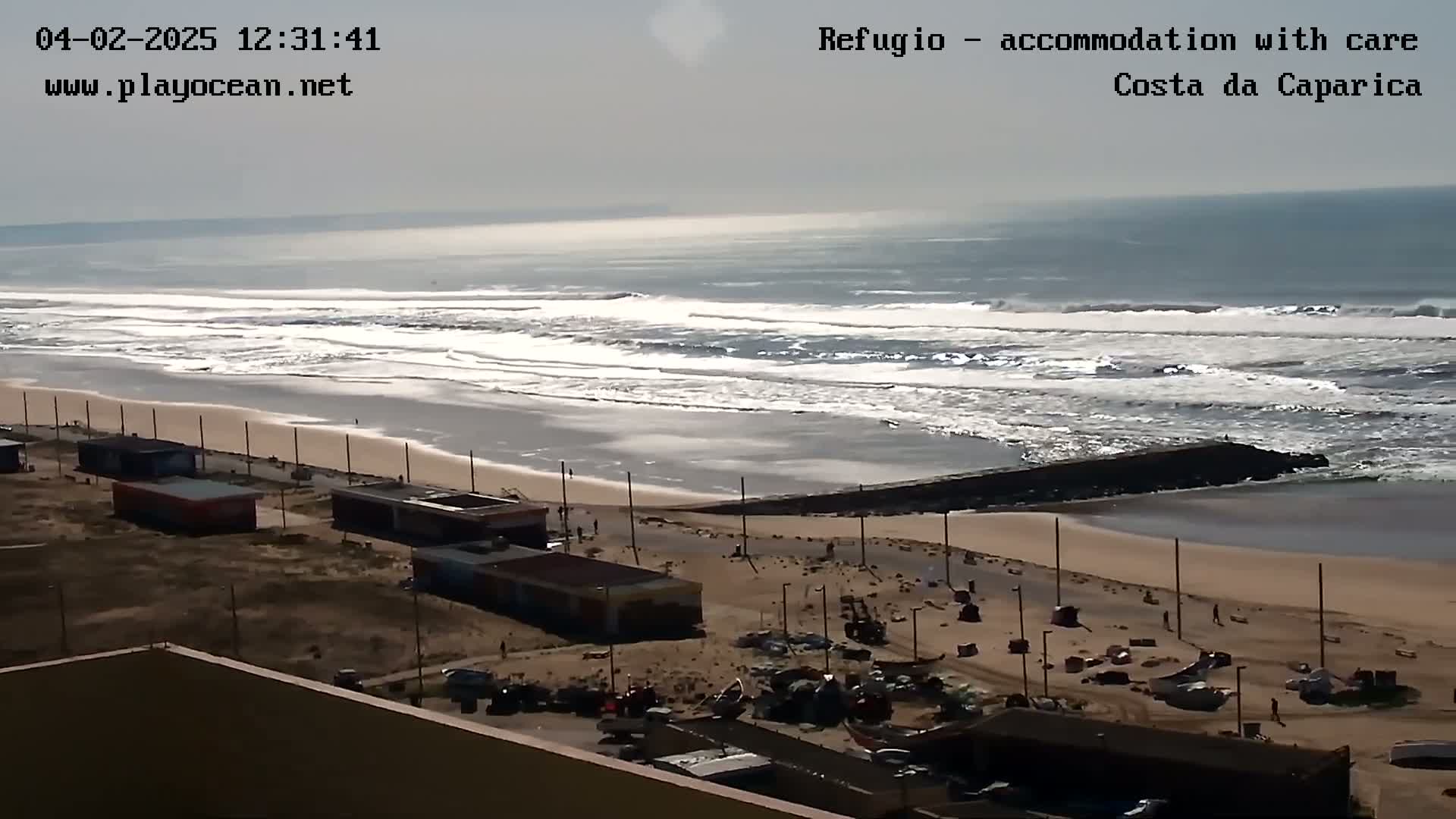 A wide shot shows a beach with moderate waves under a mostly cloudy sky, a long stretch of sand, and buildings along the shoreline.