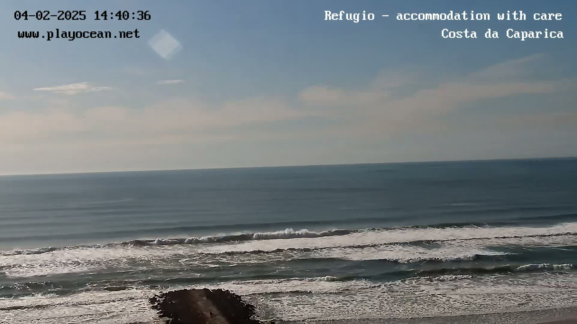 A mostly sunny day shows a view of a calm ocean with small waves breaking near the shore and a dark jetty extending into the water.