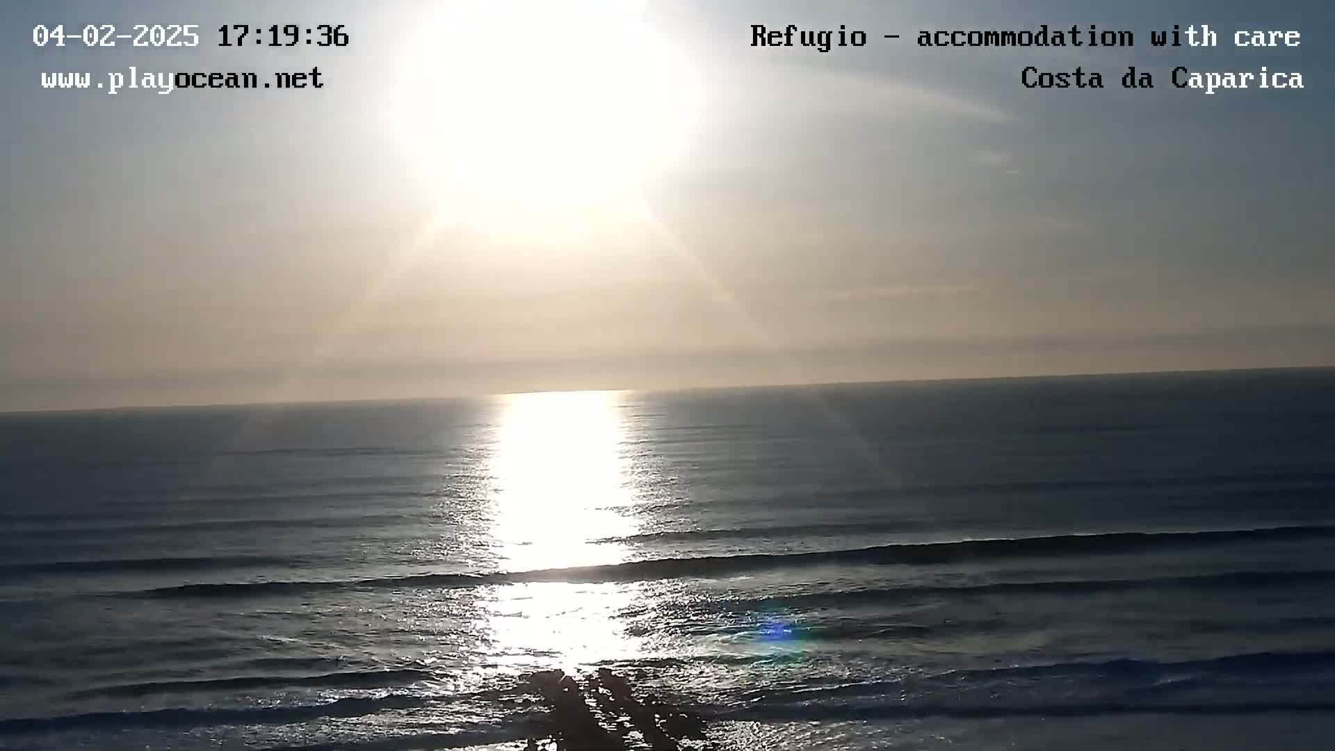 The sun shines brightly over a calm ocean with small waves rolling onto the shore under a mostly clear sky.