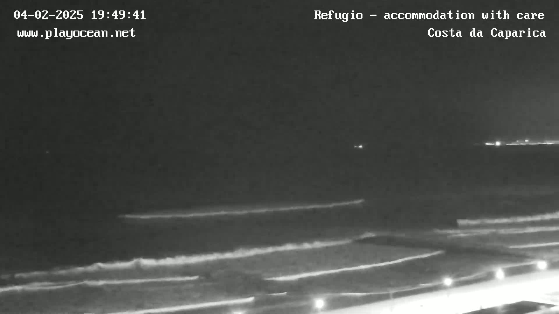 A nighttime, low-light view shows dark ocean waves lapping a shore with lights visible along the beachfront.