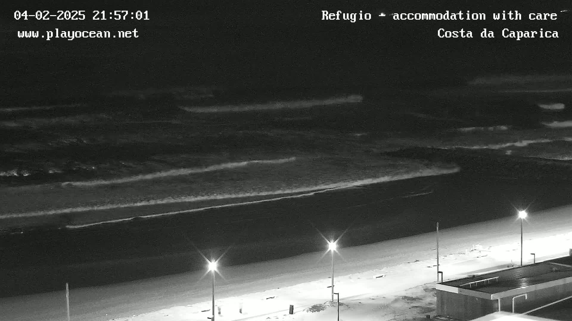 At night, dark waves roll onto a mostly empty beach under a clear sky, illuminated by a few streetlights.
