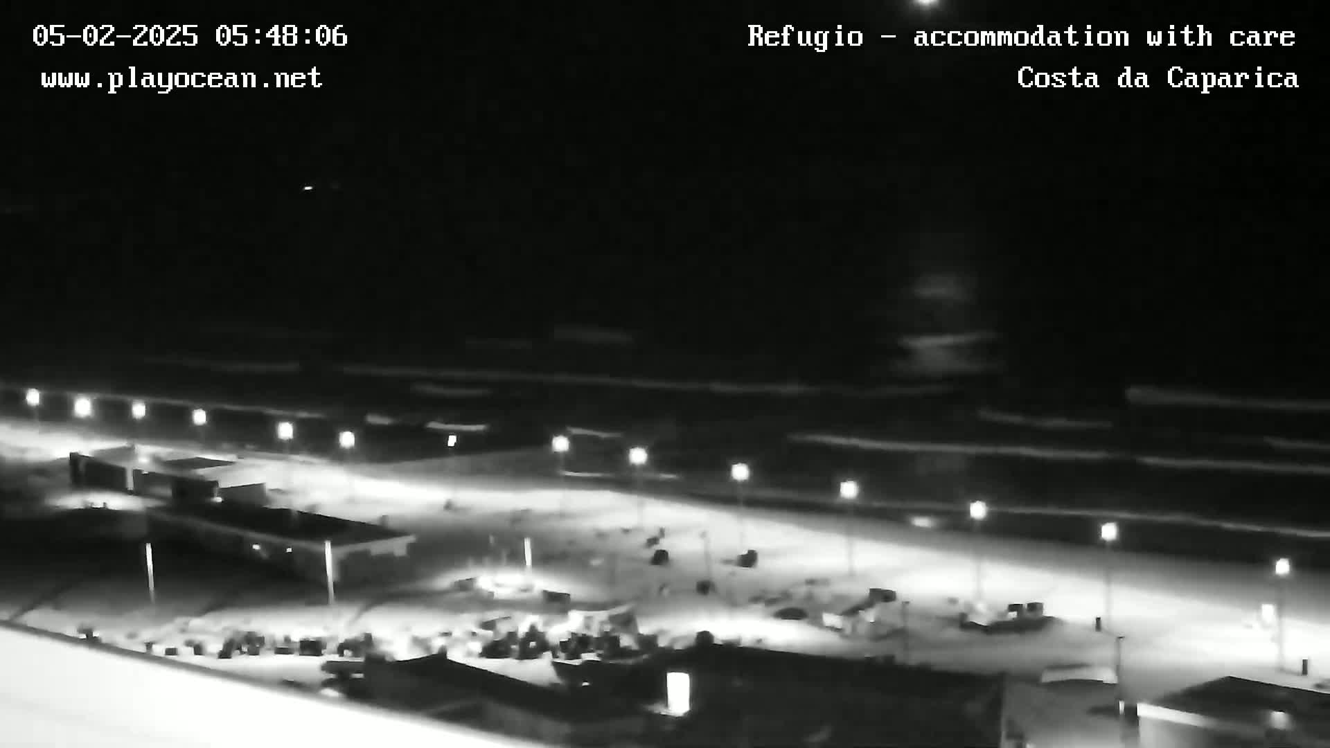 A nighttime, high-angle view shows a dark beach with waves and a line of lit-up buildings and parking areas.