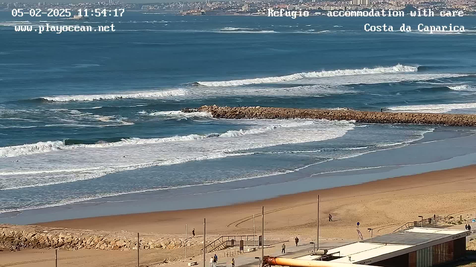 A sandy beach with a rock jetty in the ocean, showing waves breaking near the shore on a sunny day.