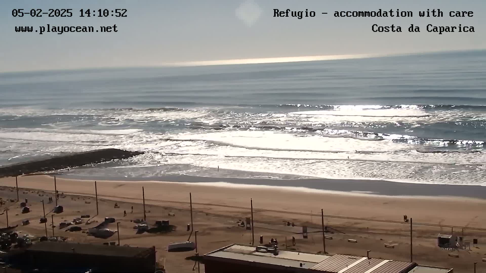 A mostly sunny day at a beach shows moderate waves breaking near a sandy shore with a few people visible on the beach and in the parking lot.