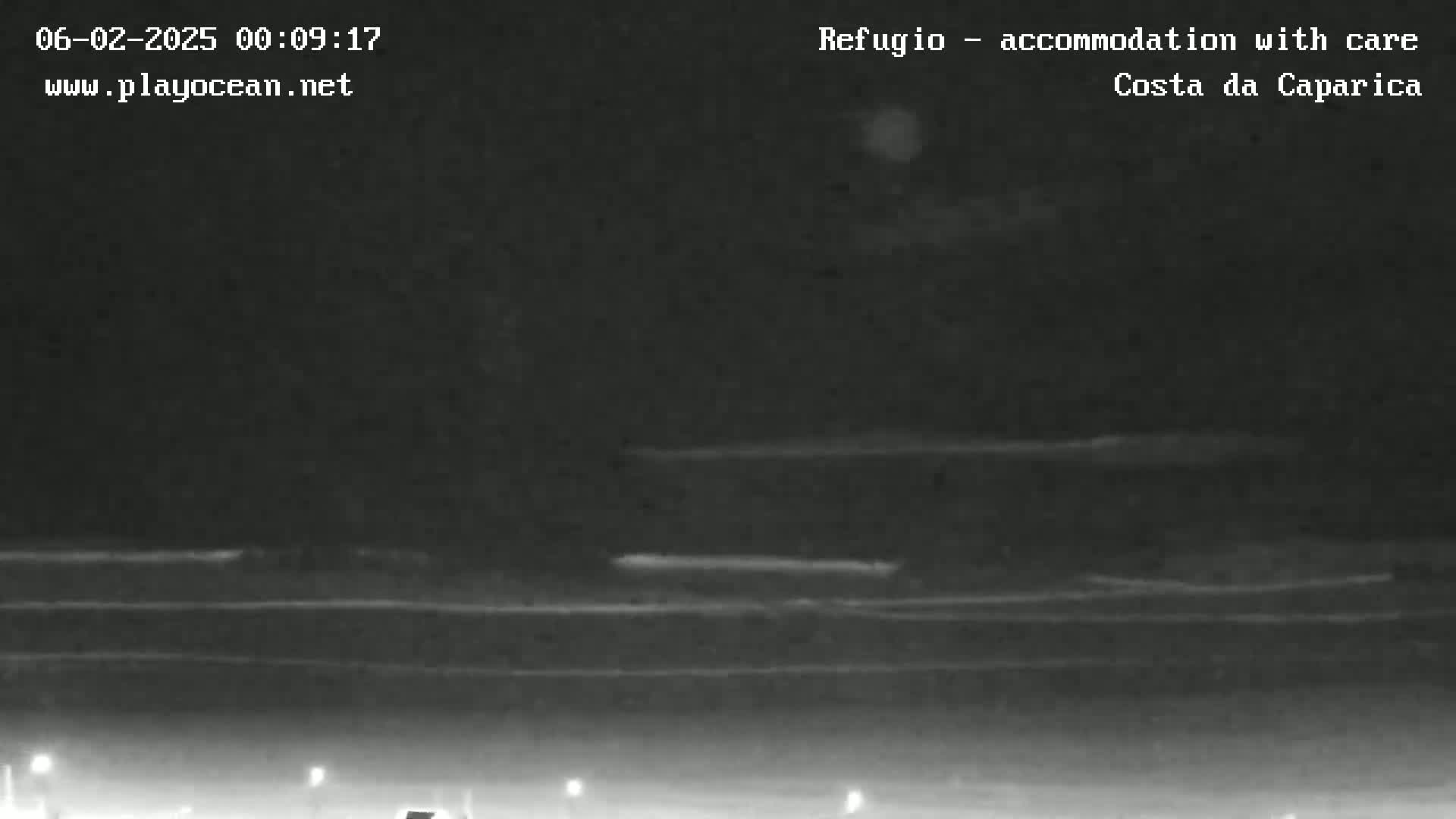 A nighttime, low-light image shows a dark beach or coastal area with faint horizontal lines suggesting waves or water, under a mostly cloudy sky.