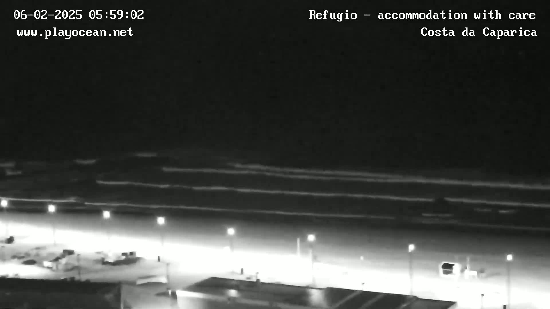 A nighttime, high-angle view shows a dark beach with gently rolling waves under a clear sky, illuminated by lights along a beachfront promenade.