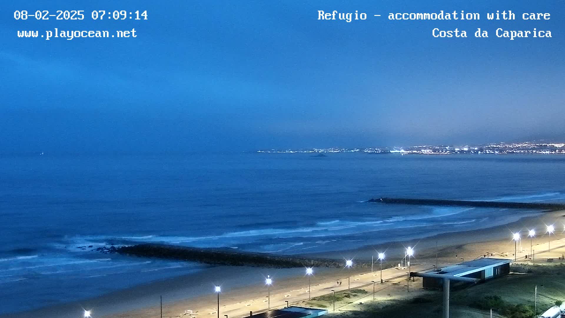 A coastal scene at twilight shows a beach with lights, a breakwater, rolling waves, and a city skyline faintly visible in the distance.