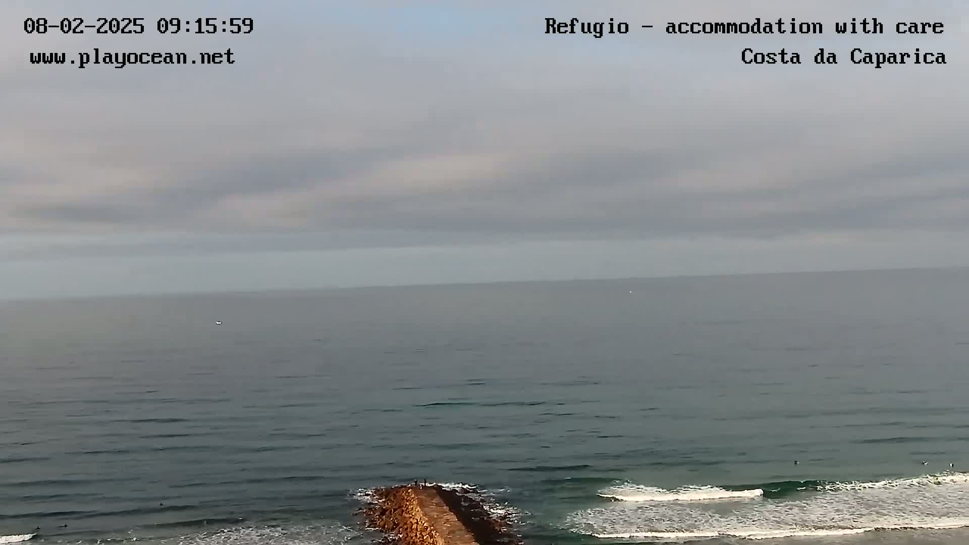 A long shot of a calm ocean with small waves breaking on a stone jetty under an overcast sky.