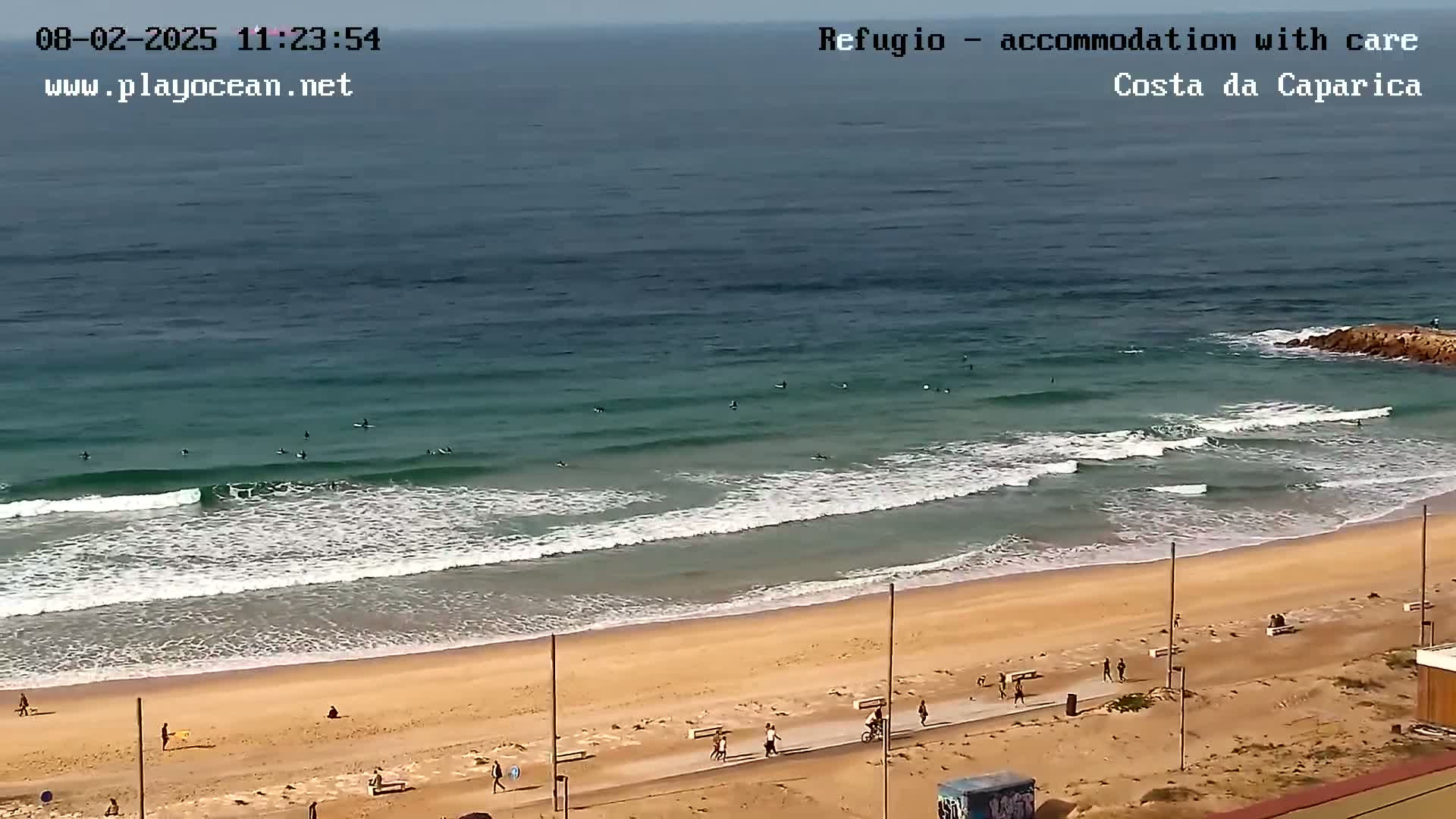 An ocean beach with gentle waves, several surfers, and a few people walking on the sand and a paved path.