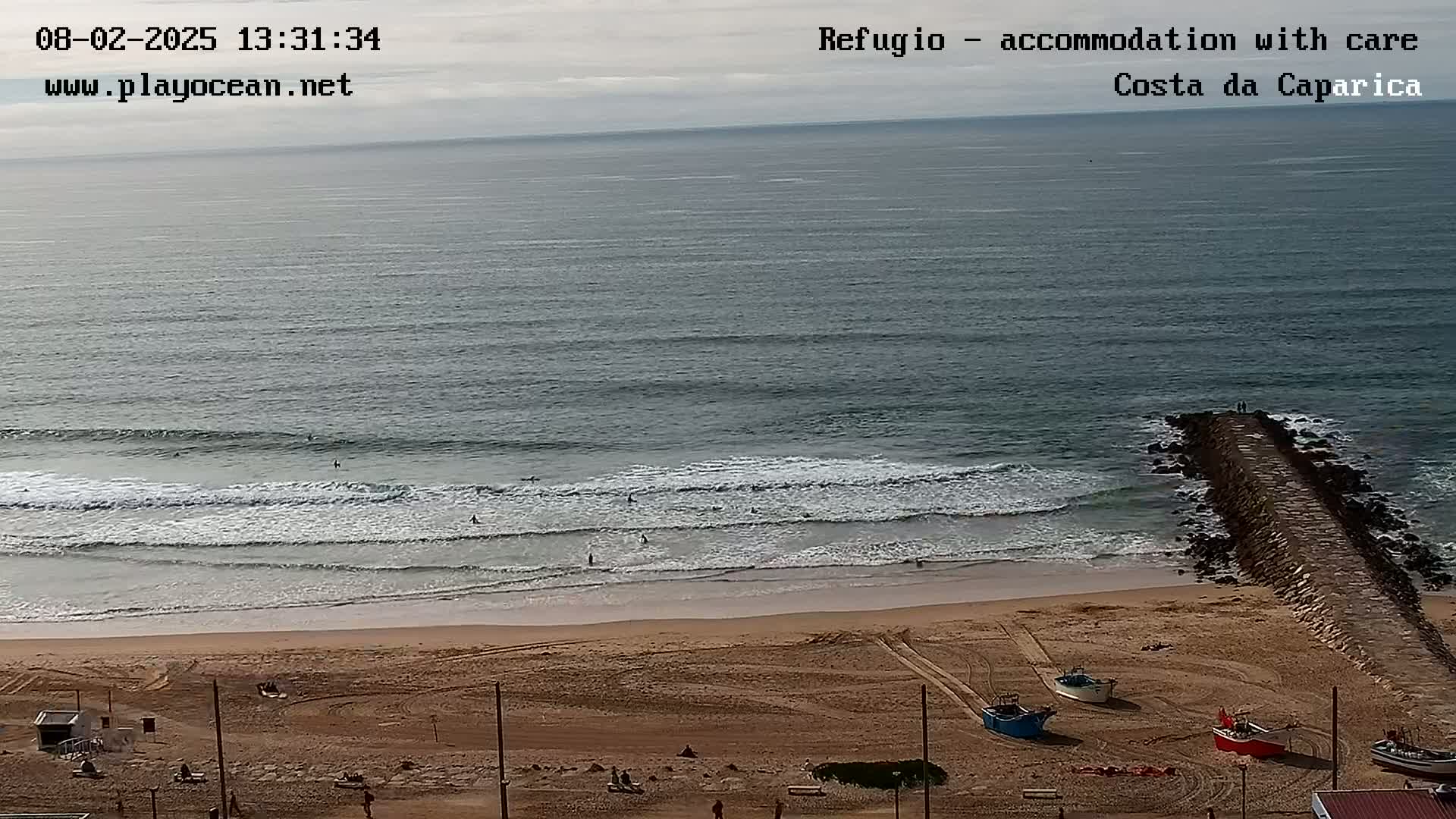 A sandy beach with several small boats, surfers in the ocean, and a long rock jetty extending into the water under an overcast sky.
