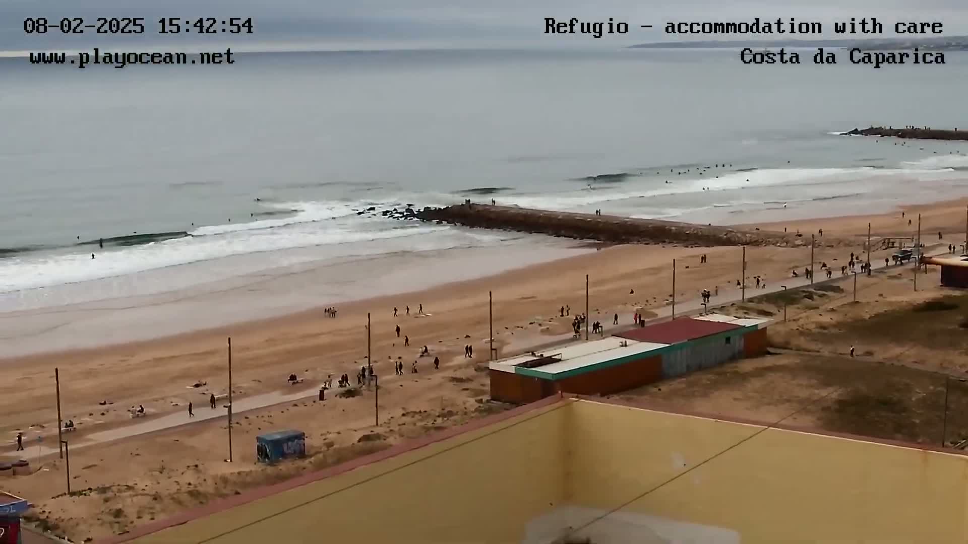 An overcast day at a sandy beach shows many people walking along the shore and in the water, near a man-made jetty extending into the ocean.