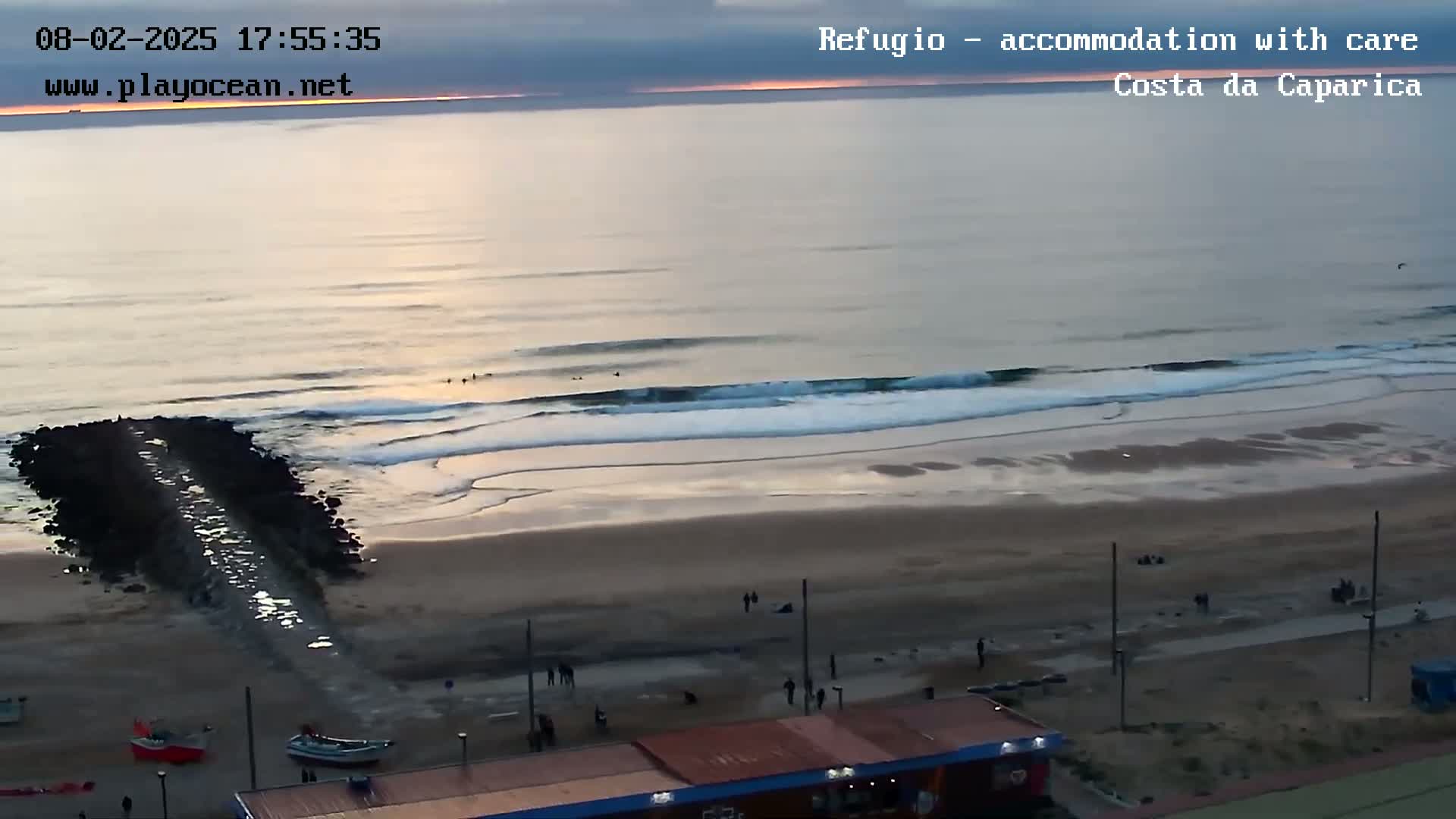 An ocean beach at sunset with gentle waves, a few people on the sand and in the water, and a rock jetty extending into the water.