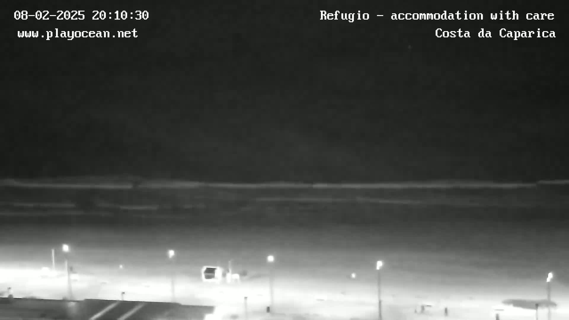 A nighttime, low-light view shows a dark beach and ocean with a few lights visible along the shoreline.