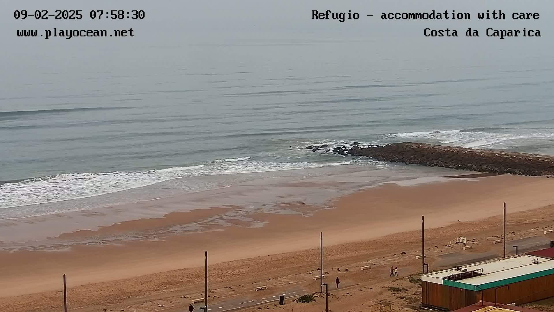 A sandy beach with a rock jetty is visible, with gentle waves rolling in under an overcast sky.