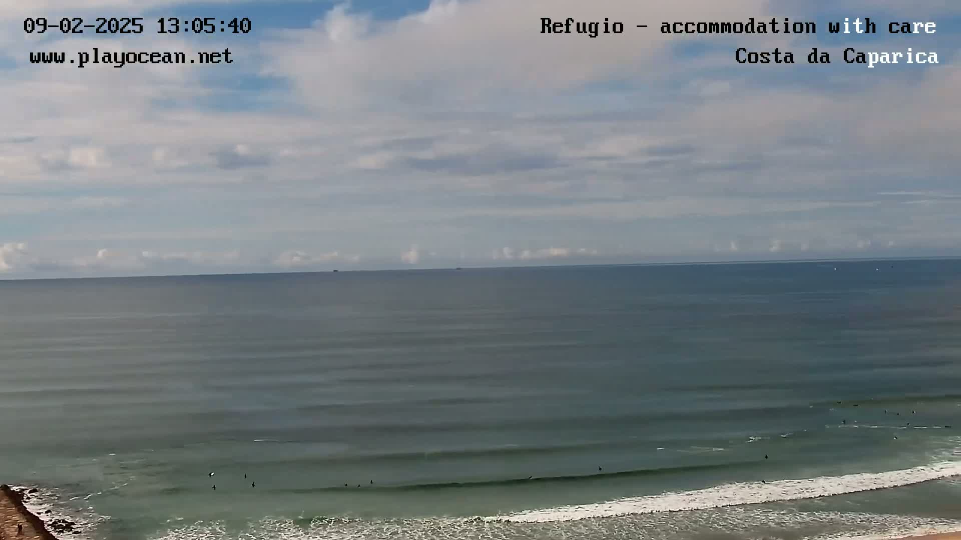 A mostly cloudy sky overlooks a calm ocean with several surfers in the water near the shore.