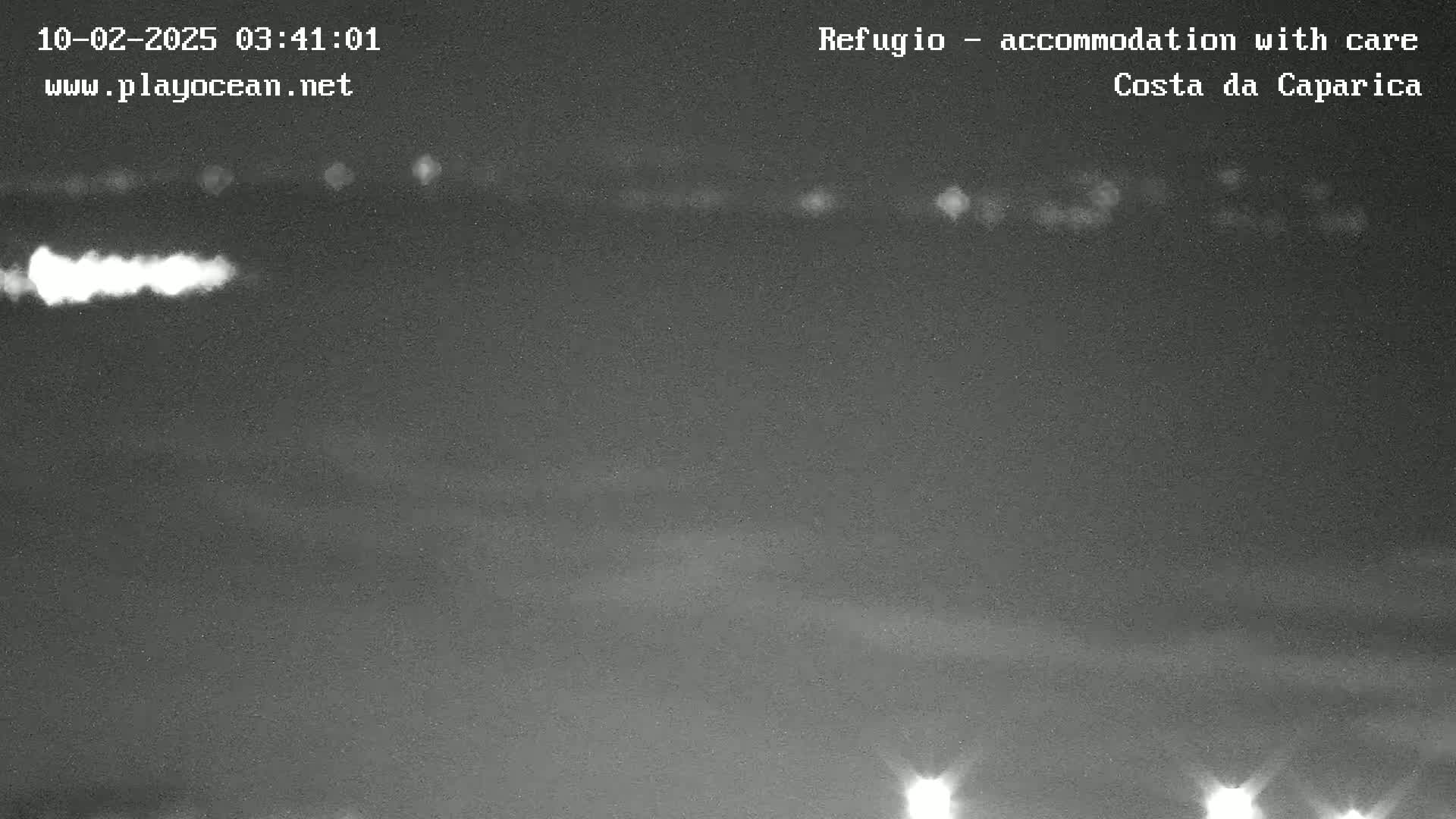 A nighttime, low-light image shows a dark body of water with faint lights on the horizon and a brighter light source in the lower left.