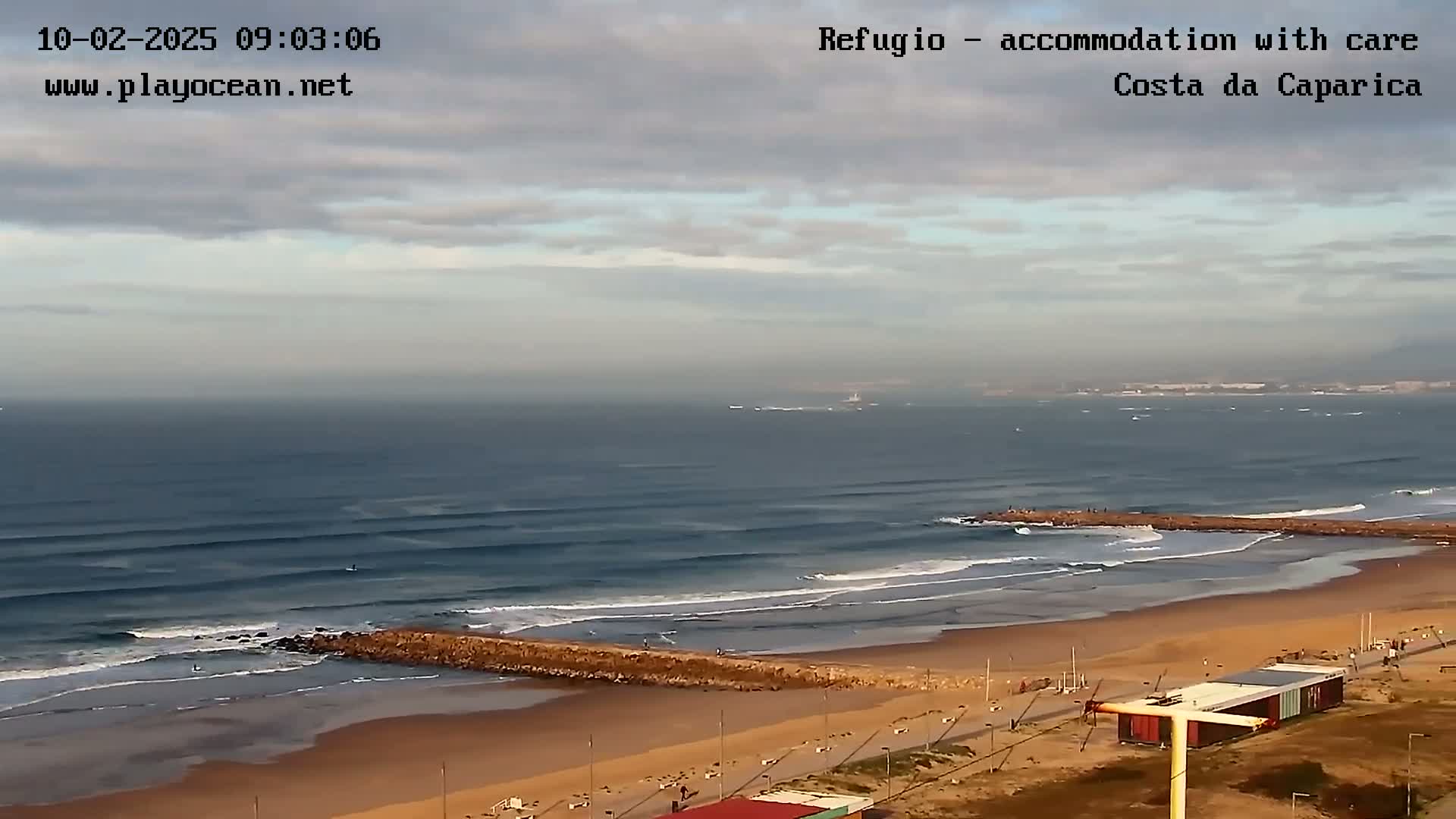 An expansive view of an ocean beach on a partly cloudy day shows waves breaking near a long jetty, with a sandy shoreline and some buildings visible in the distance.