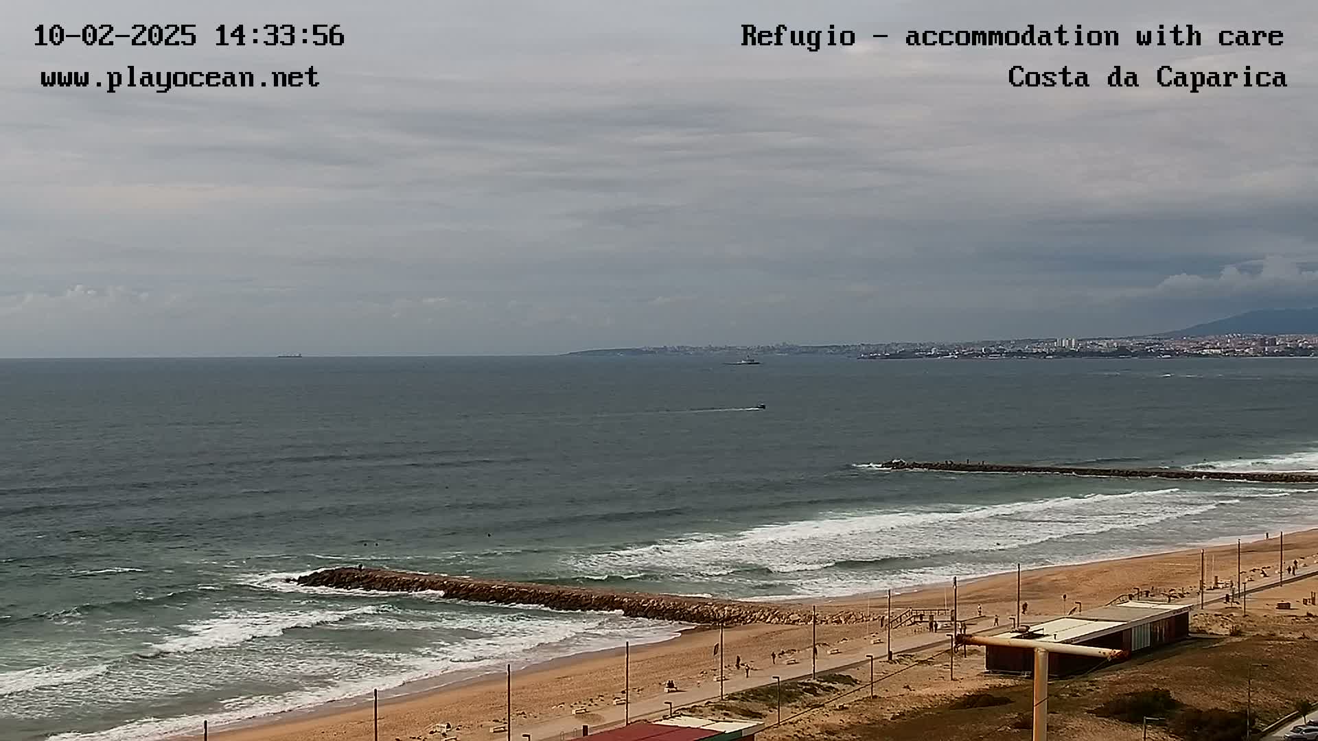 A long sandy beach with a wave breaker extends to a calm ocean under a cloudy sky.