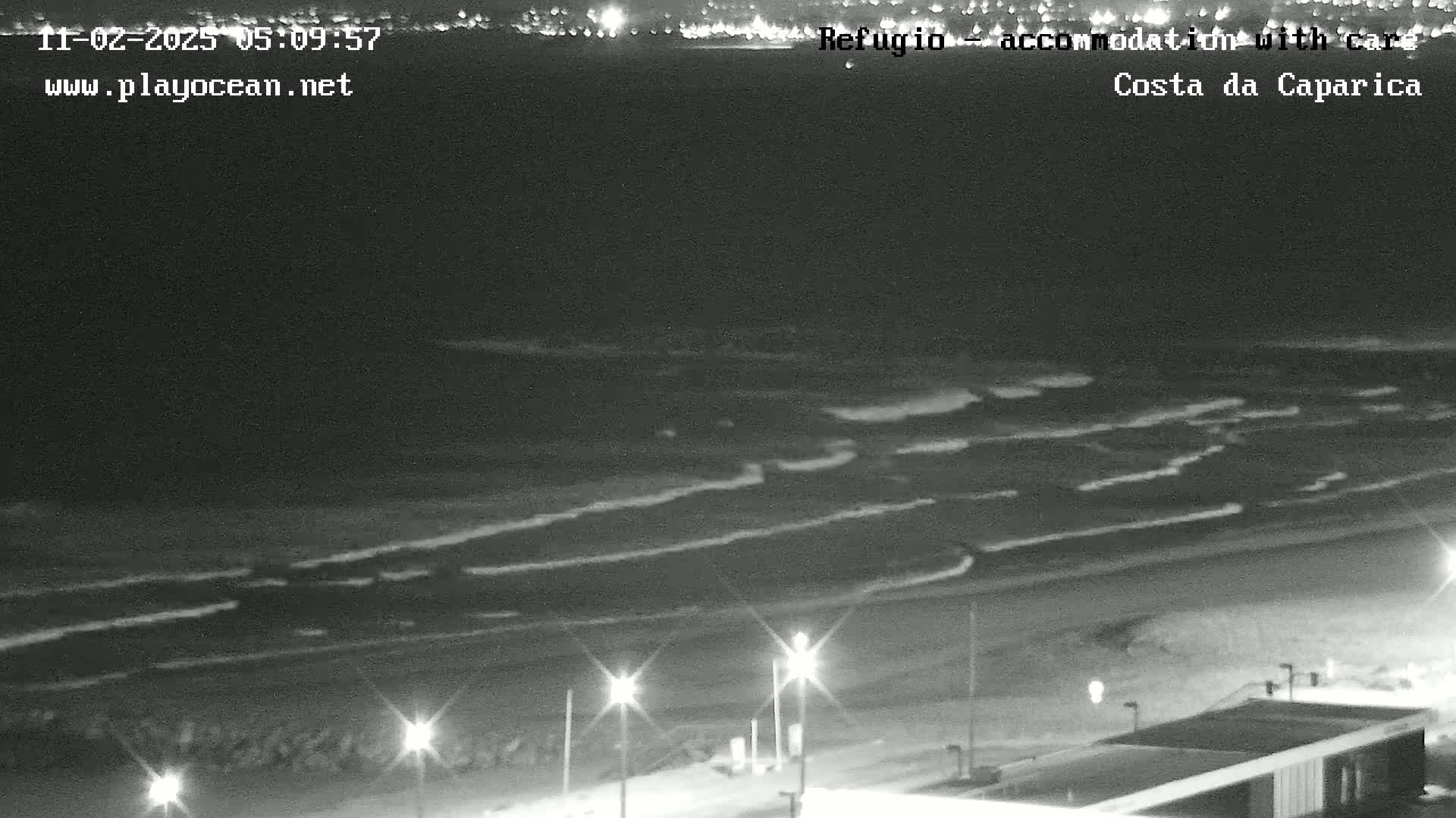 A nighttime, black-and-white view of a beach with gentle waves lapping the shore, illuminated by streetlights.
