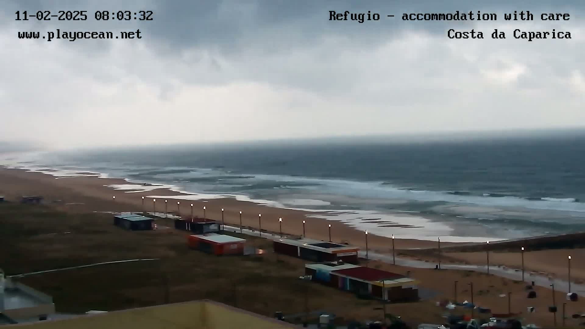 An overcast sky hangs over a beach with moderately rough waves, several small buildings line a walkway along the shore.