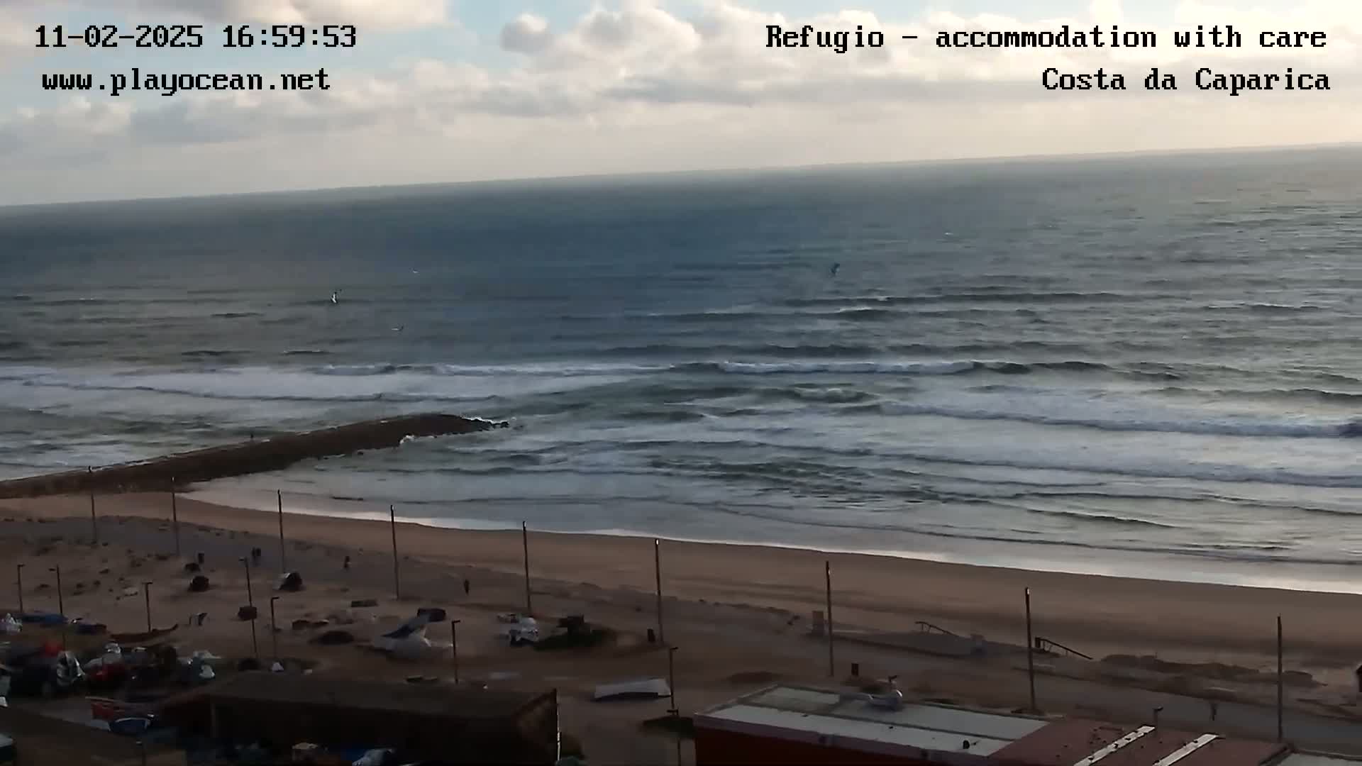 A mostly cloudy day shows a beach with a rocky jetty extending into a dark-blue ocean with small waves rolling onto the shore.