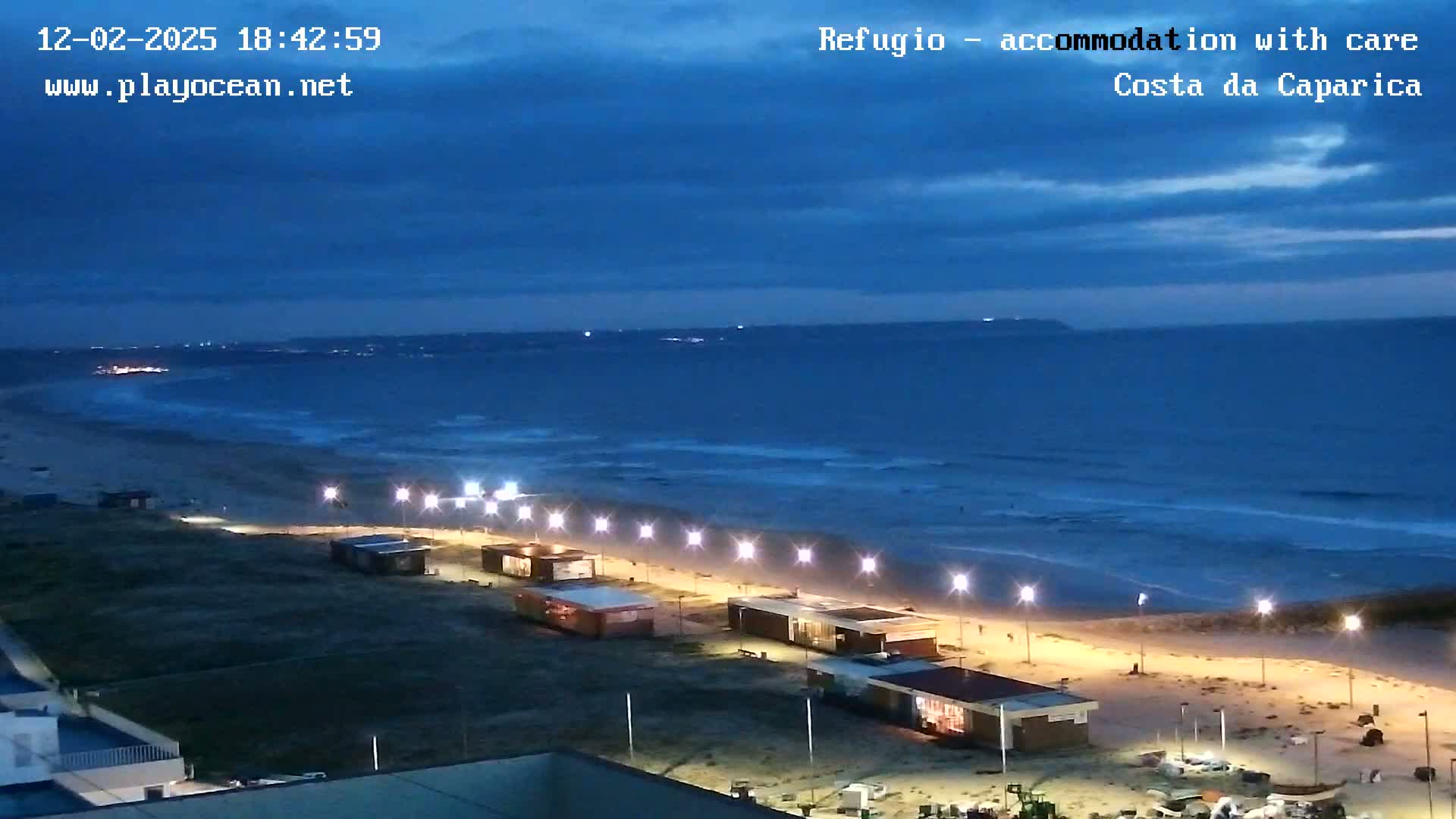 A dark-blue ocean with waves rolls onto a sandy beach at dusk, where a line of illuminated buildings and lampposts stretches along the shore.