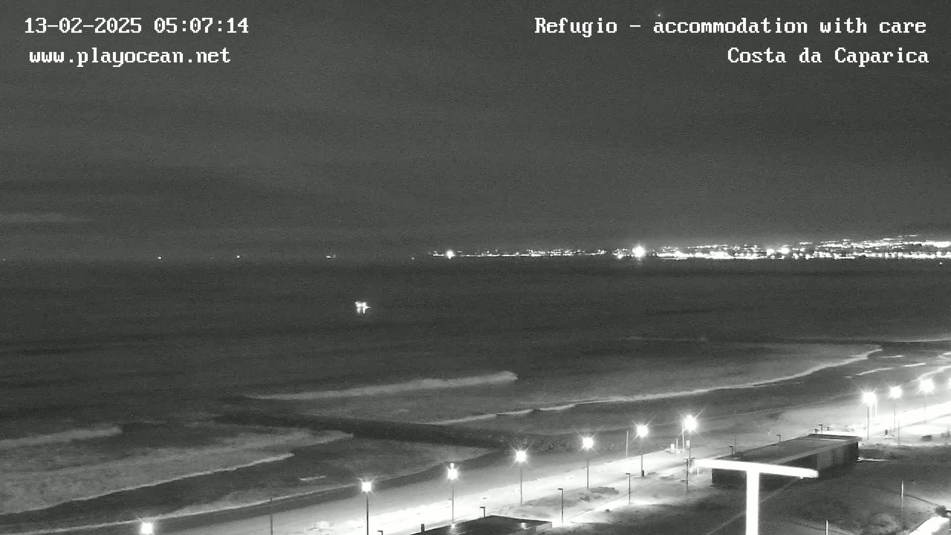 A nighttime, black-and-white view of a beach with gentle waves lapping the shore, illuminated by streetlights, and a city skyline faintly visible in the distance.