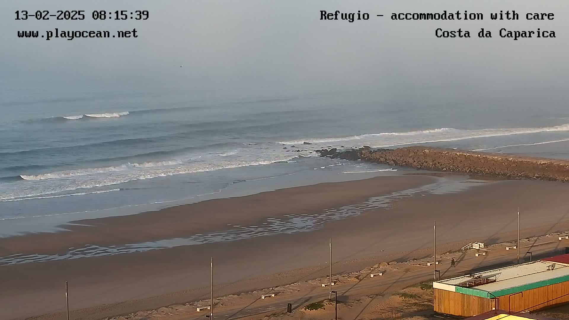 A hazy, overcast day reveals a sandy beach with small waves rolling onto shore, meeting a rocky jetty.