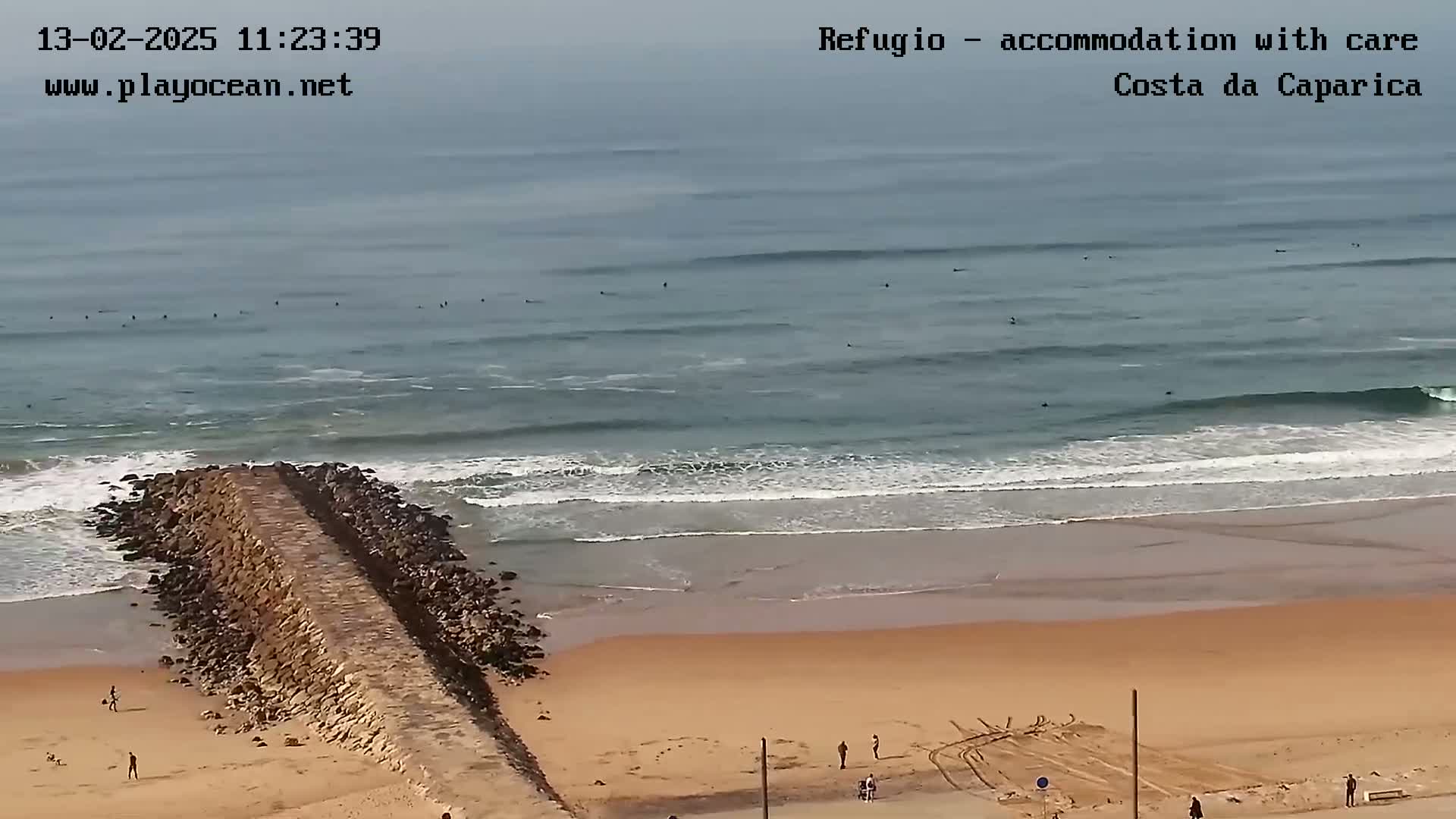 A long, rocky jetty extends from a sandy beach into a somewhat overcast ocean where several surfers are visible.