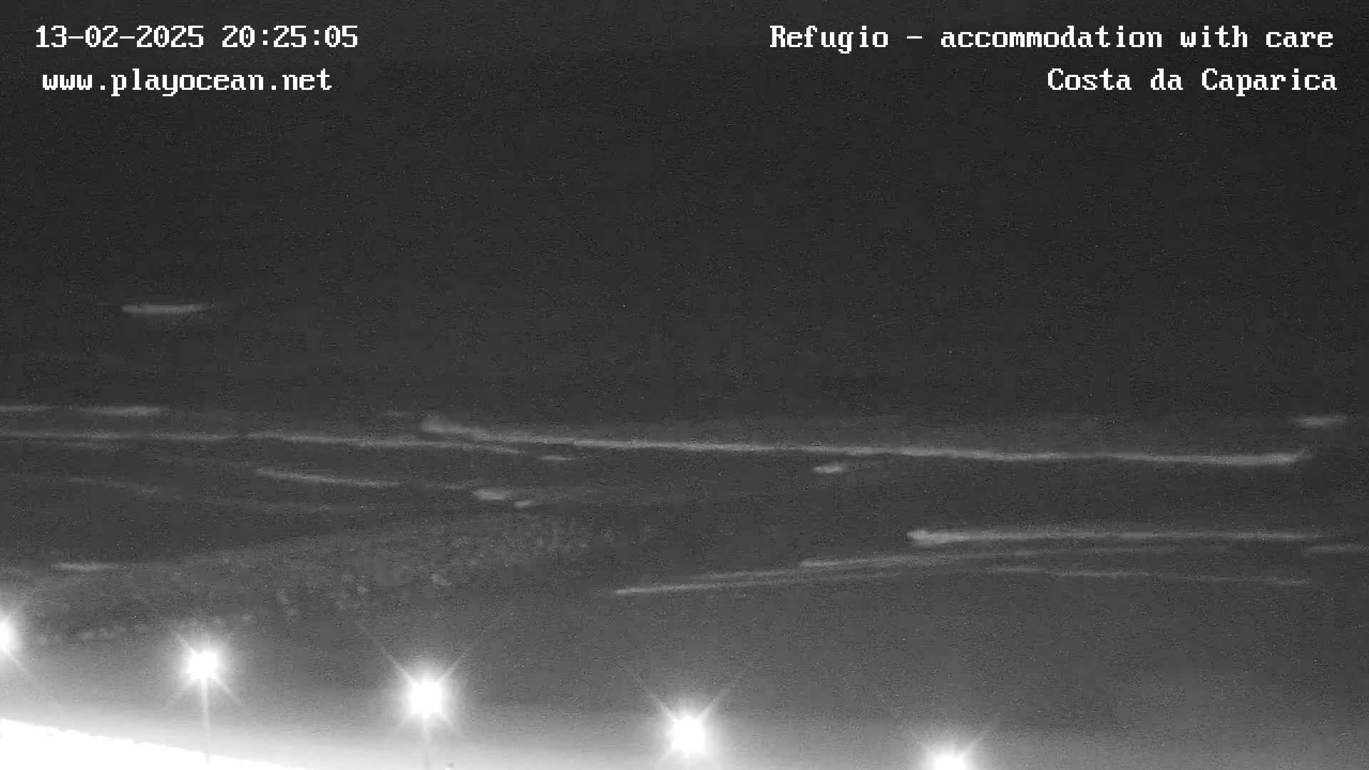 A nighttime, low-light image shows a dark ocean with subtle waves under a mostly clear sky, illuminated by several lights in the foreground.