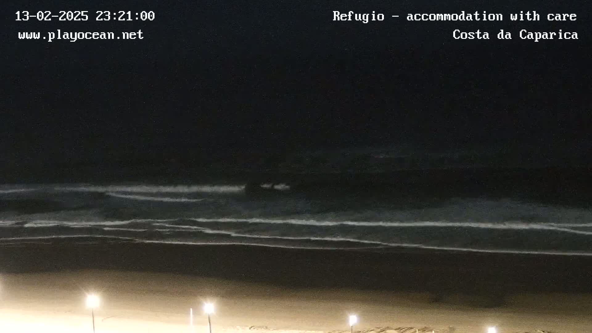 At night, gentle waves lap a dark sandy beach under a clear, dark sky illuminated by a few lights.