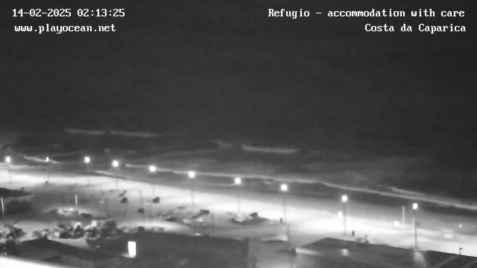 A nighttime, low-light view shows a beach with gently rolling waves under a dark sky, illuminated by a line of streetlights along a roadway behind it.