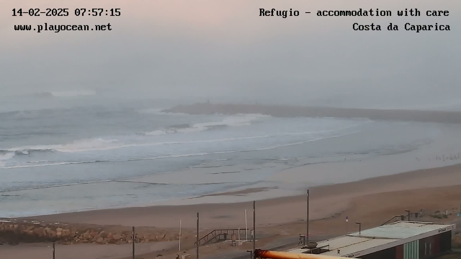 A hazy, overcast day reveals a beach with moderate waves rolling onto the shore, meeting a long sandy expanse that leads to a paved area with buildings.