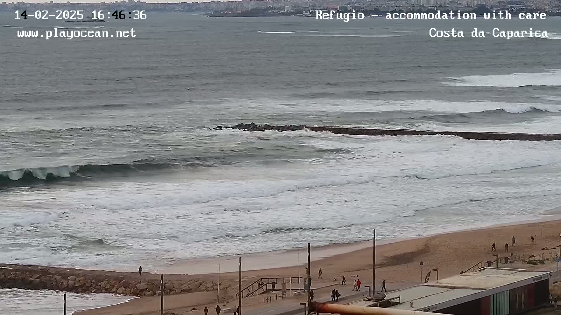 A mostly cloudy day shows a beach with moderate waves and a few people walking along the shore.