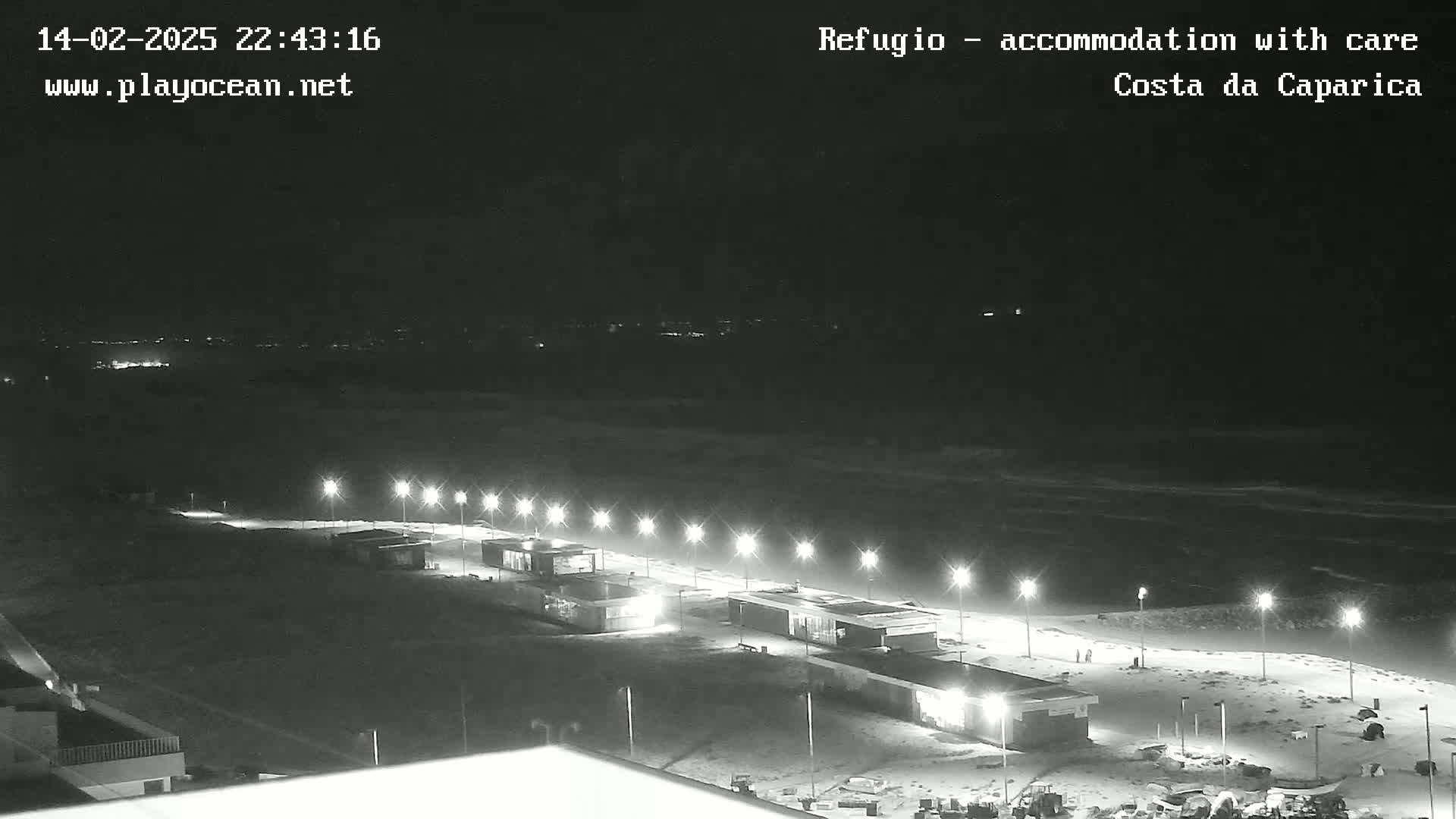 A nighttime, high-angle view shows a dark, ocean beach with a row of lit structures along the shoreline under a clear sky.