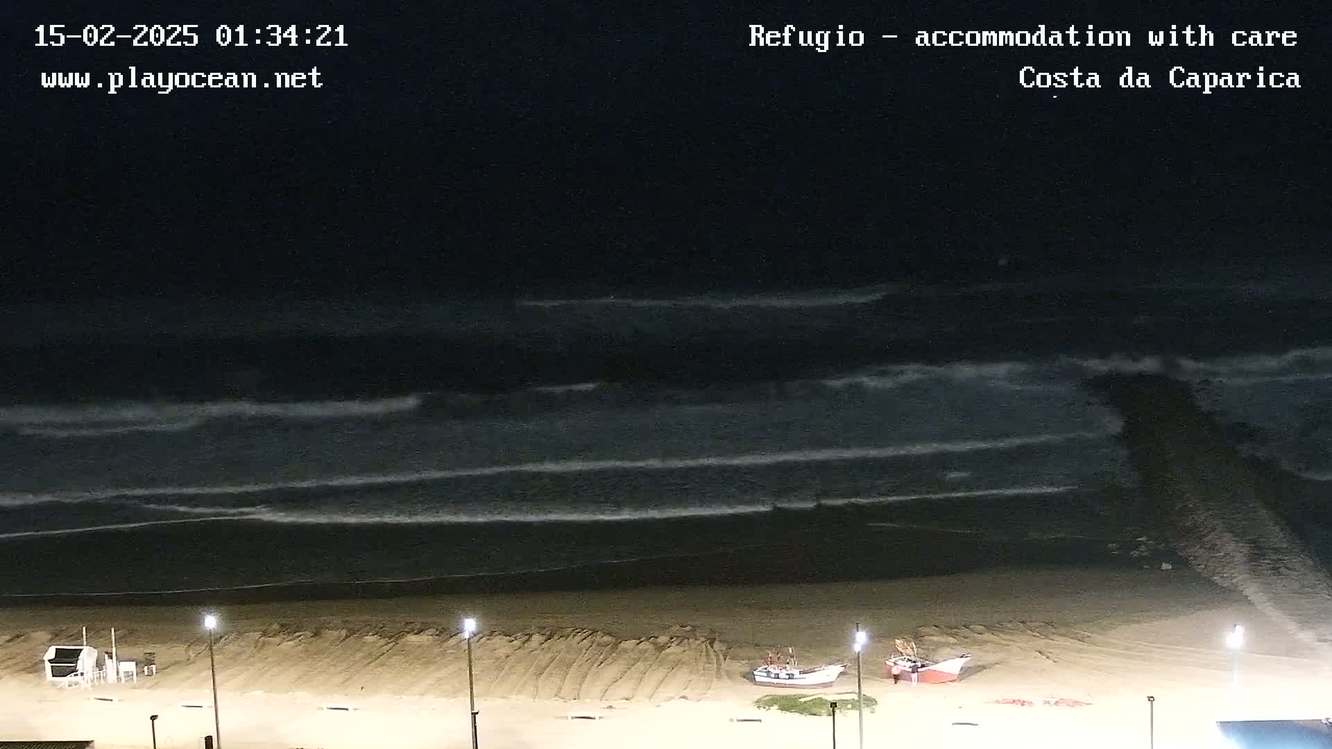 At night, several waves roll onto a sandy beach where two small boats are beached near lit lampposts.