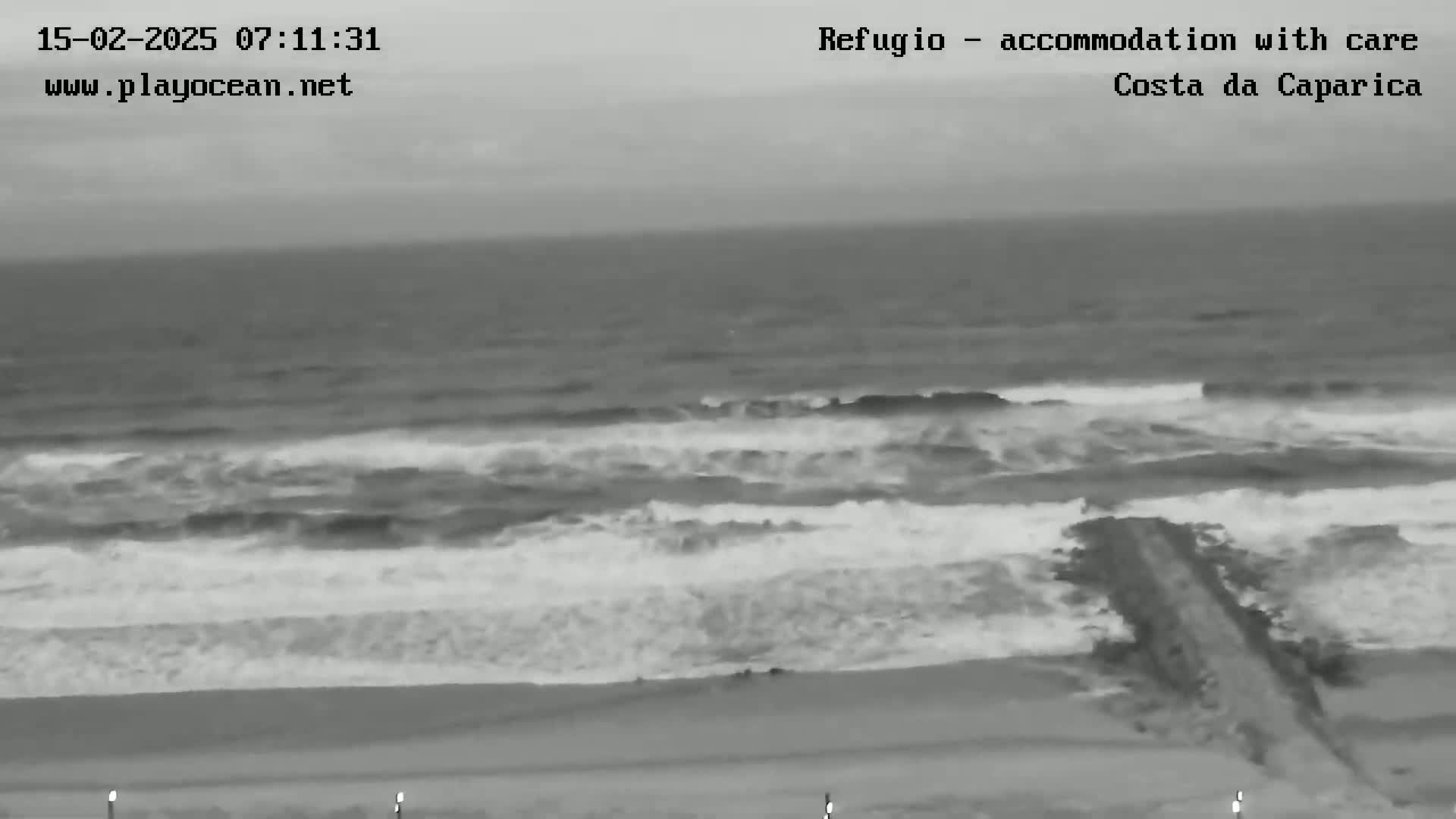 A grayscale image shows a beach with moderate waves crashing on the shore under an overcast sky.