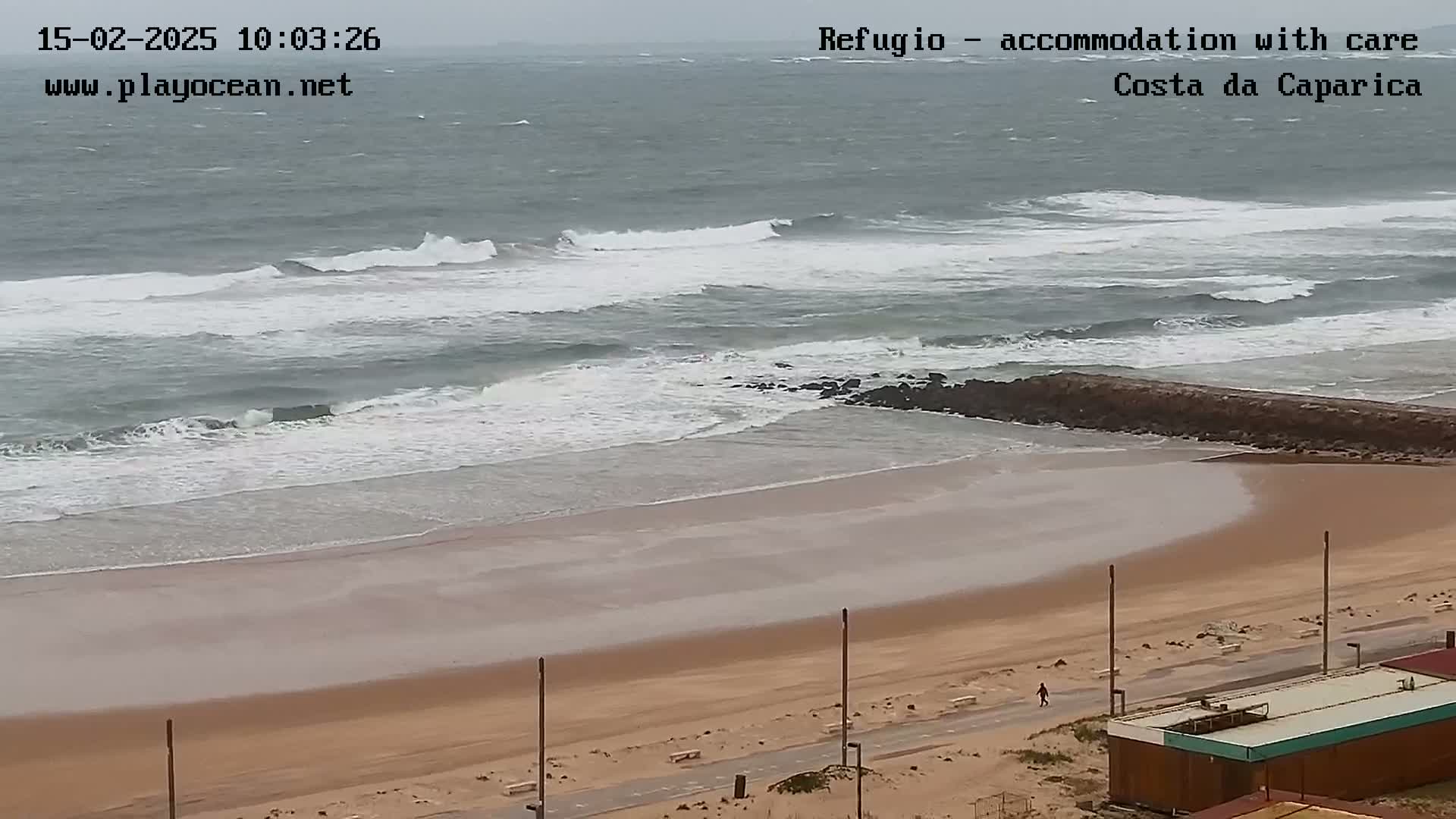 A sandy beach with moderate waves under an overcast sky is seen from above, with a person walking along the shore and buildings in the lower right.