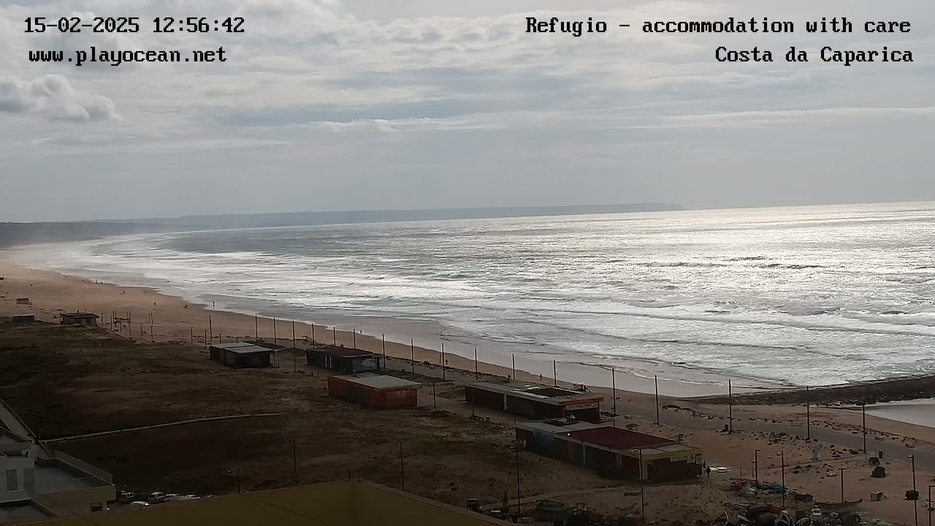A sandy beach with small buildings and a road runs alongside a wavy ocean under a cloudy sky.