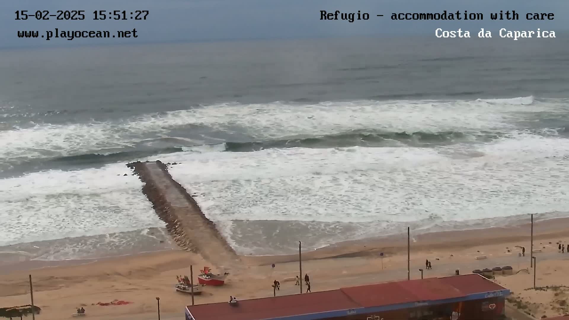 A rocky jetty extends into a choppy ocean with white-capped waves crashing on a sandy beach, where a few people are walking near a building.